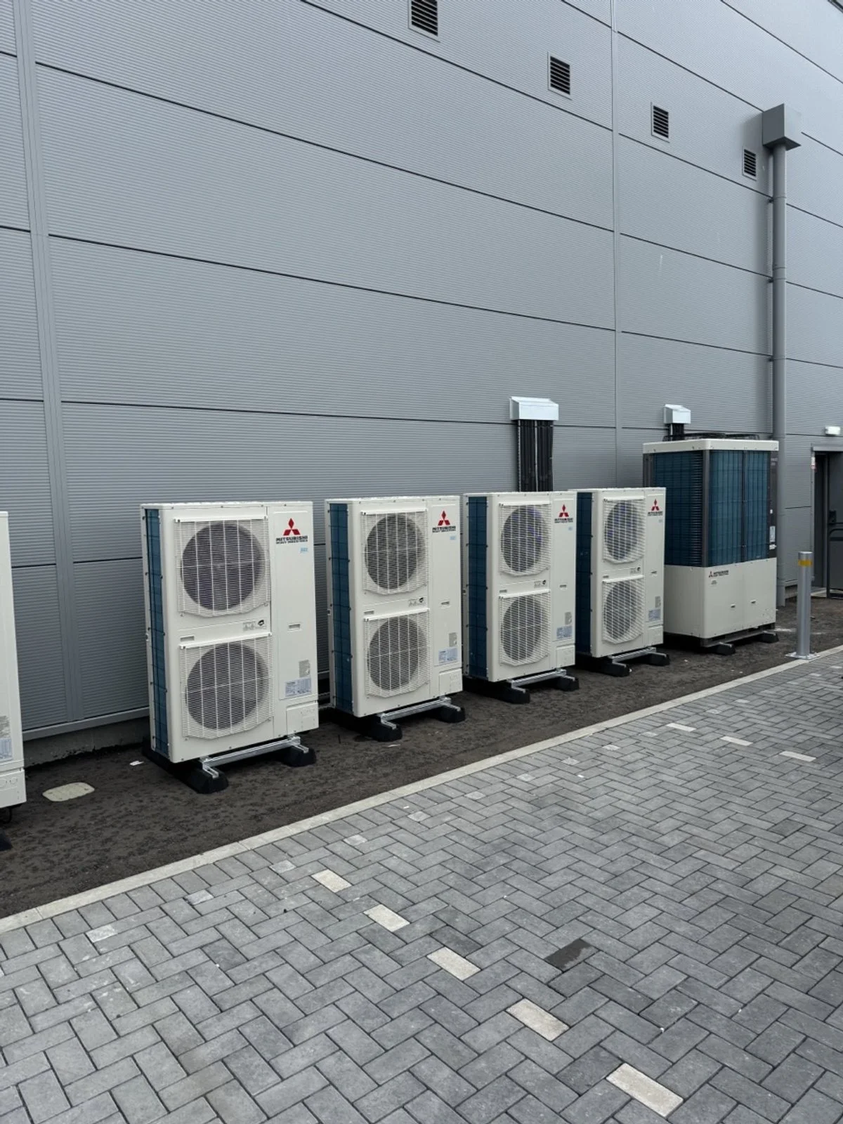 Multiple outdoor air conditioning units installed against a gray building wall, with a paved sidewalk in front.