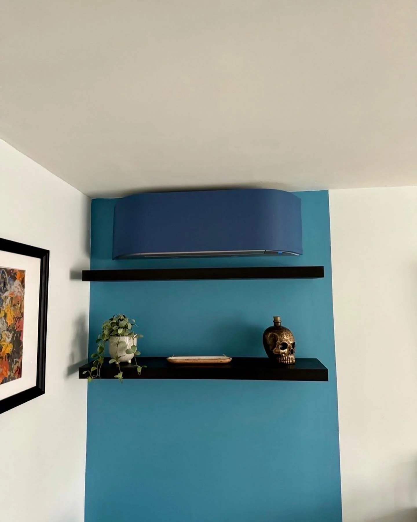 Decorative wall with two black shelves, a potted plant, a wooden tray, and a skull figurine, against a blue accent wall with a blue fabric light fixture above.
