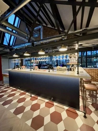 Modern bar area with a white countertop, hanging pendant lights, and a tiled floor with a geometric pattern, inside a stylish industrial-style space with exposed beams and large windows.