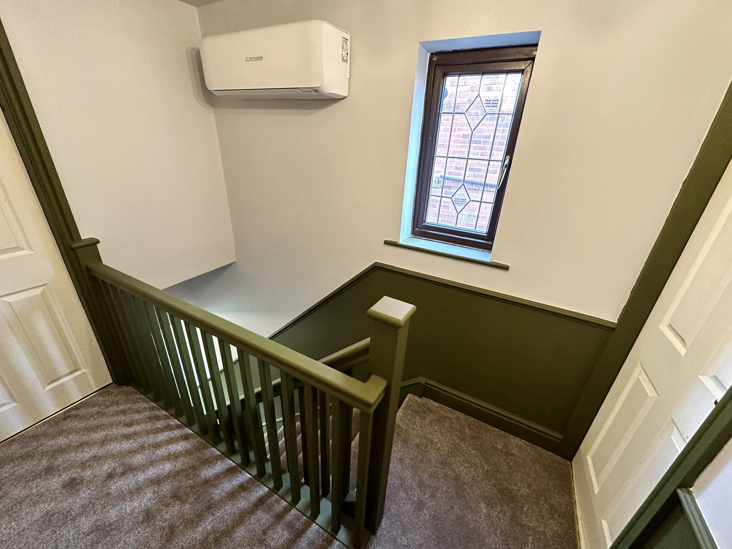Indoor staircase landing with green railing, beige carpet, window with metal grill, white wall, and air conditioning unit.