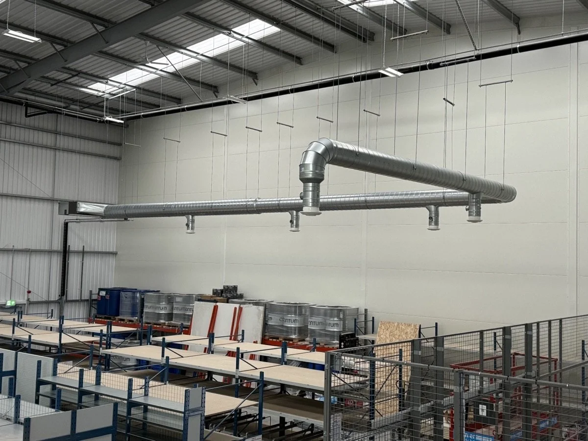 An industrial warehouse with metal ductwork hanging from the ceiling, along with work tables, storage containers, and shelving.
