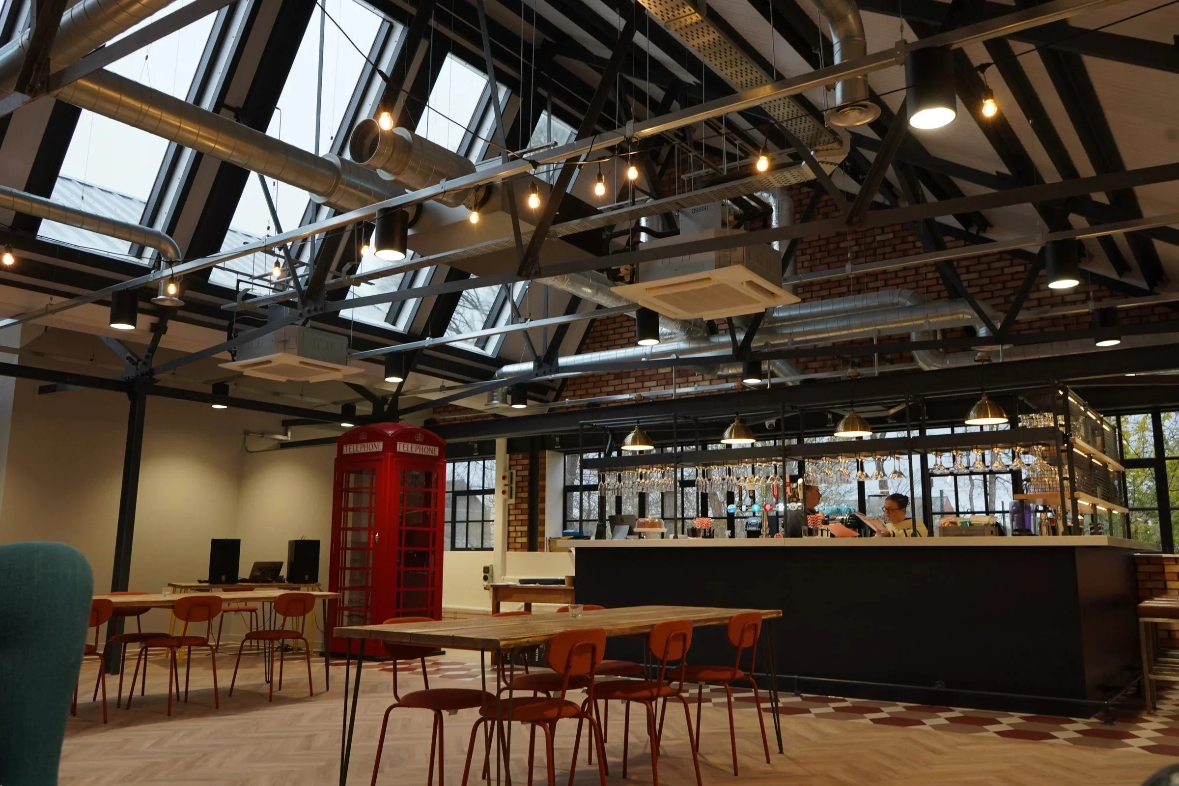 Interior of a modern café with a high ceiling featuring exposed ductwork and large skylights. There are tables and chairs, a red telephone booth, and a barista preparing drinks behind the bar.