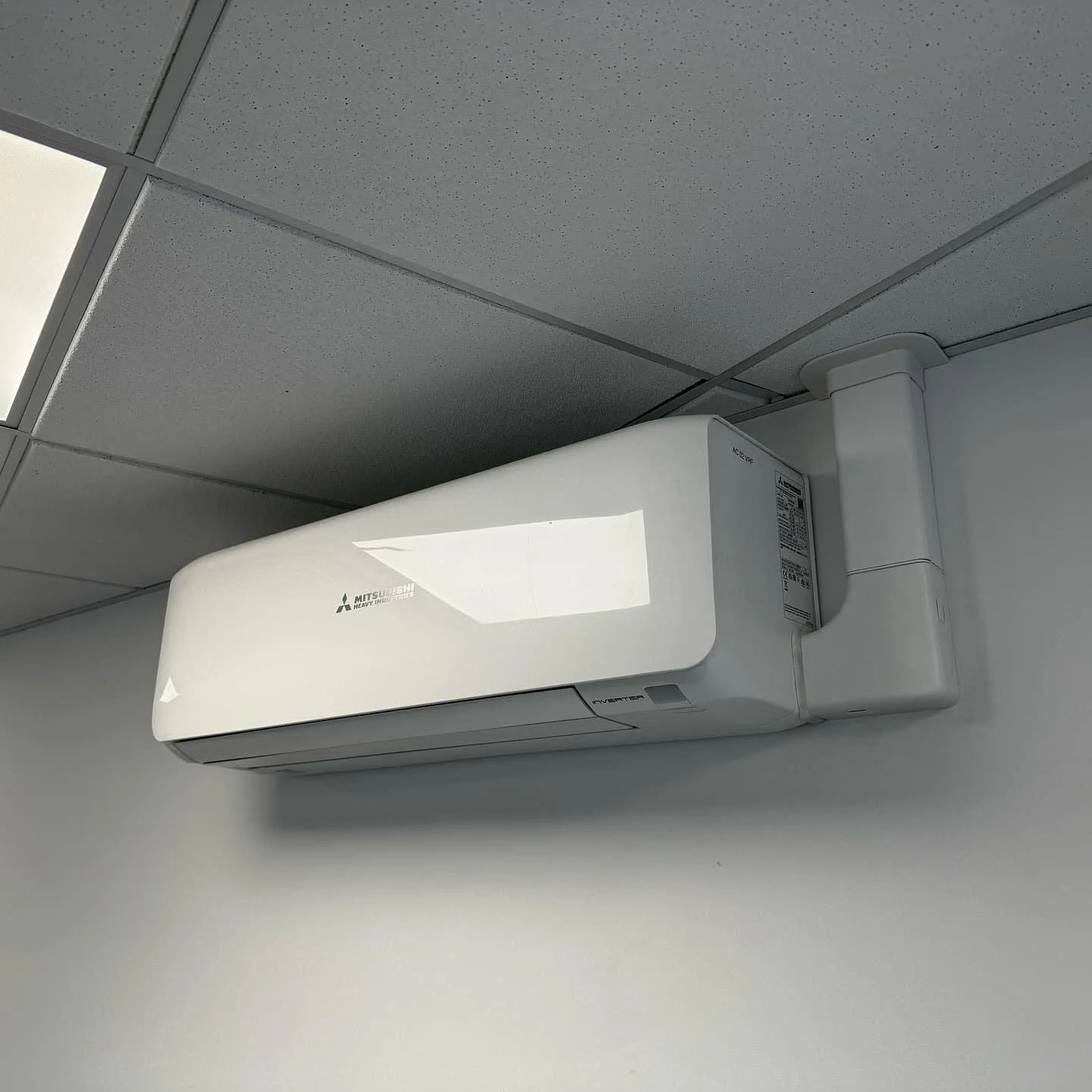 A Mitsubishi heavy industries wall-mounted air conditioner unit installed on a white wall near a ceiling with gray acoustic tiles.