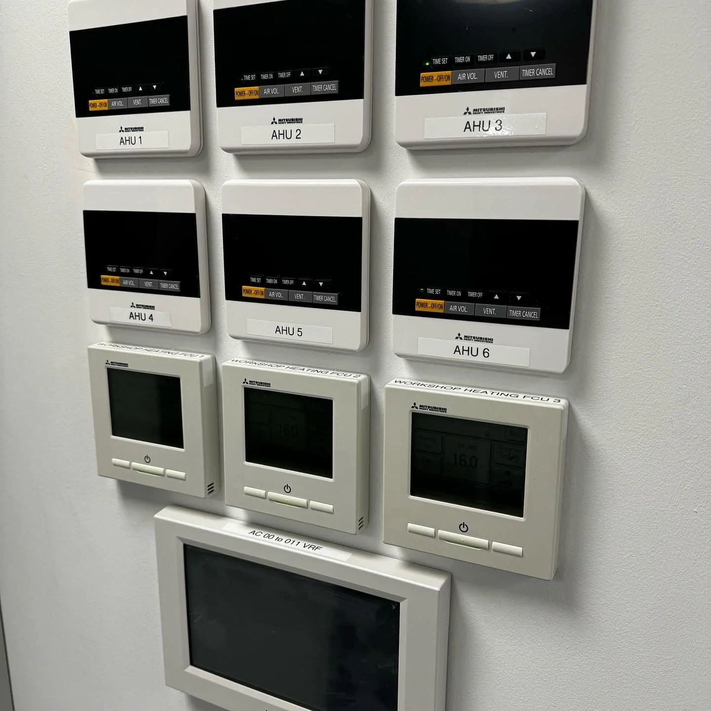 A wall with multiple control panels, including six black Mitsubishi units labeled AHU 1 to 6, and three white digital thermostats, with the lower one showing a temperature of 16.0.