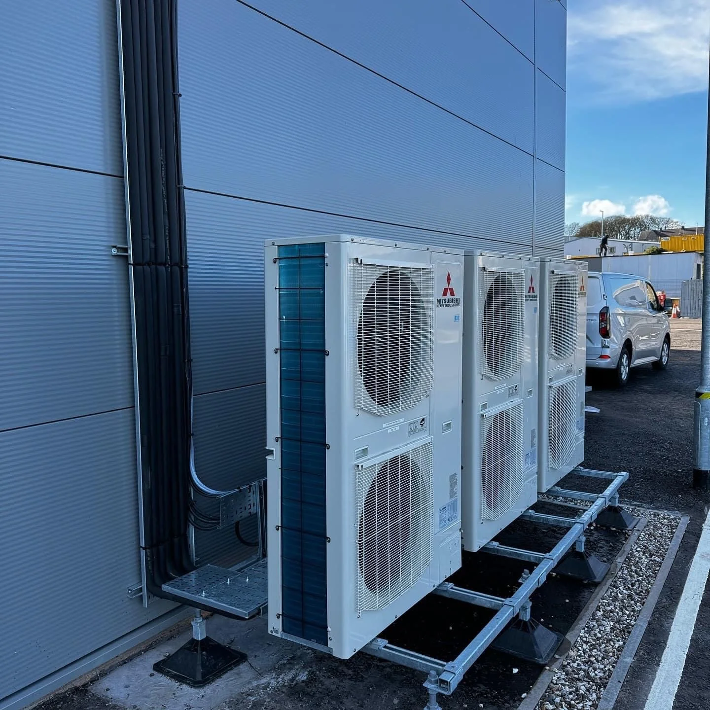 Four Mitsubishi HVAC units installed outside on a metal platform near a metallic building wall, with a white van and other buildings in the background.