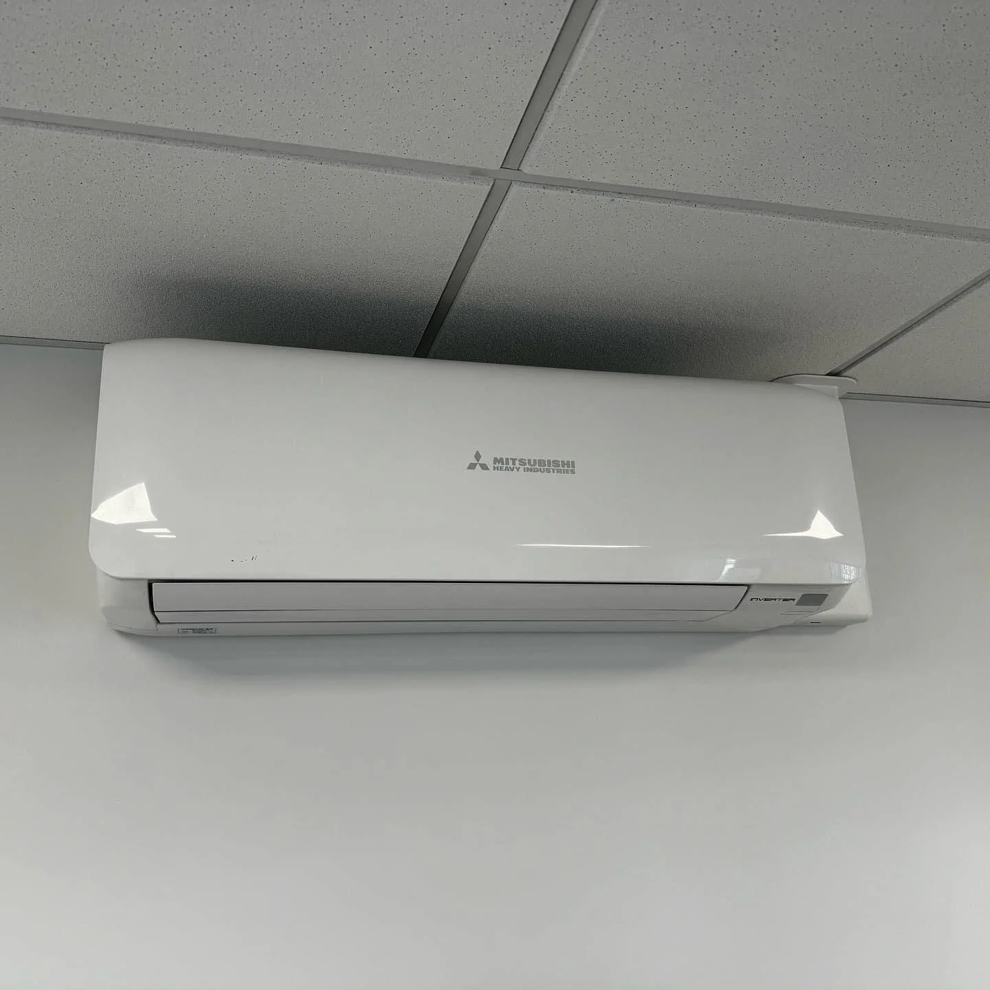 White Mitsubishi Heavy Industries wall-mounted air conditioner on a white wall, with a gray ceiling above.