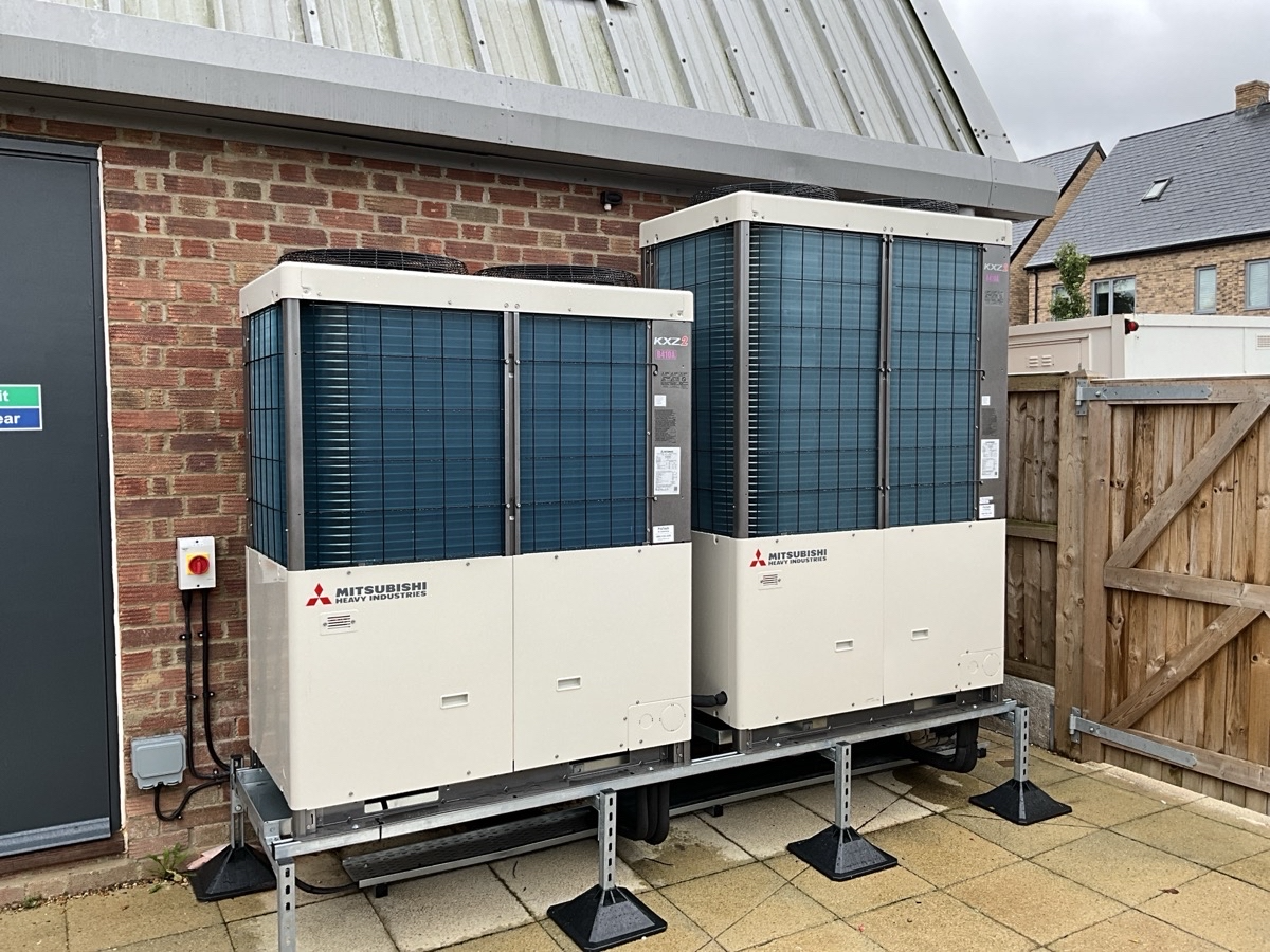 Two Mitsubishi air conditioning units outdoors next to a brick building, elevated on a metal platform with adjustable feet.