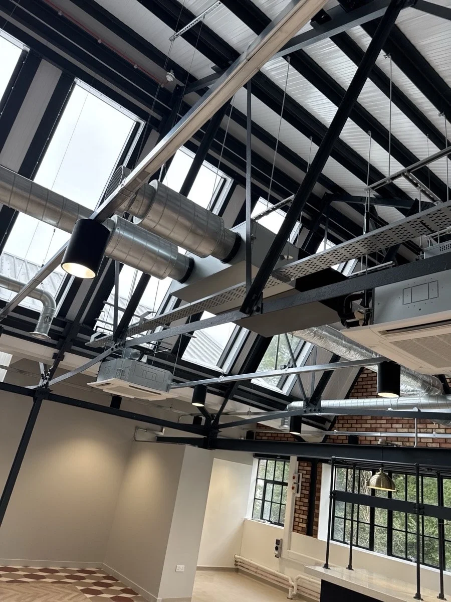 Interior view of an industrial-style building showing exposed ductwork, large windows, and a high ceiling with metal beams and skylights.