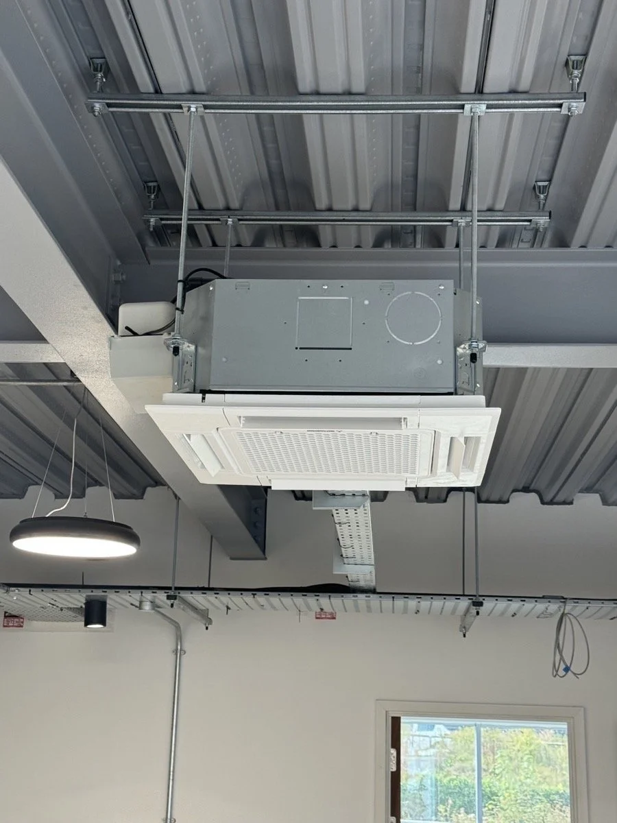 Ceiling-mounted HVAC duct and vent system with exposed metal framework and ceiling grid, a circular light fixture, and a window in the background showing outside greenery.