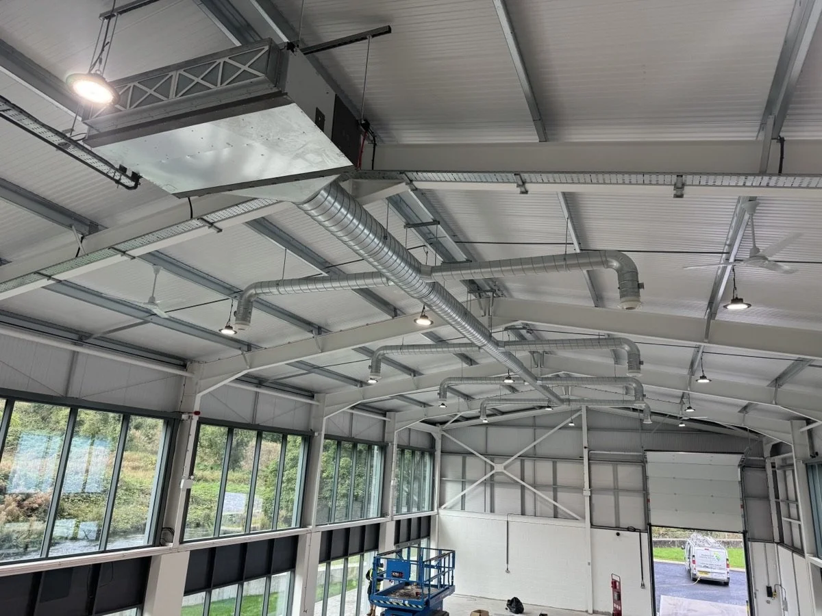 Interior view of a large industrial or warehouse space with high ceilings, metal ductwork, large windows, and a partially open roll-up door.