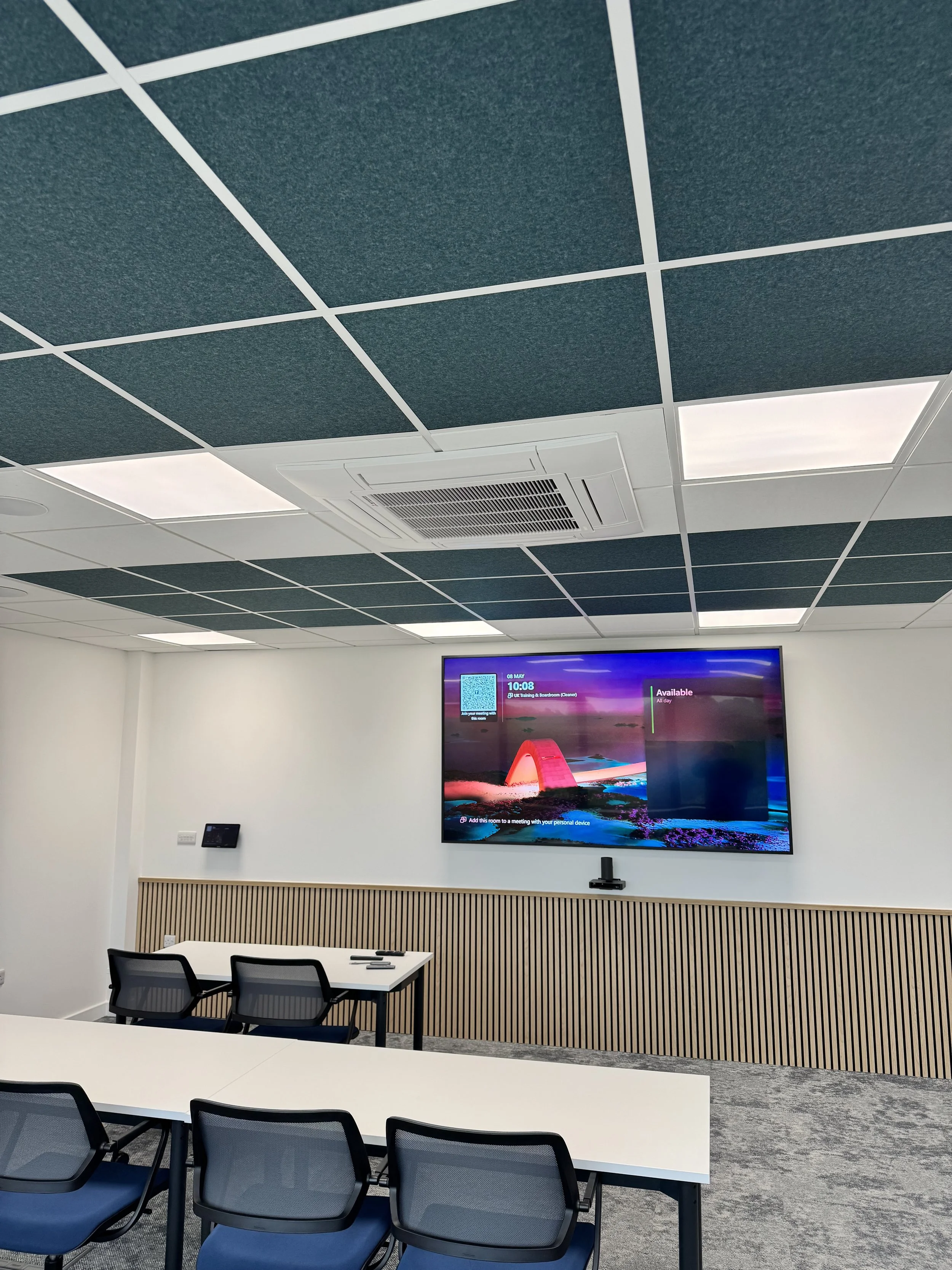 Modern conference room with white walls, blue acoustic ceiling panels, a large monitor displaying a landscape image, a wooden slatted panel underneath, and chairs around tables.
