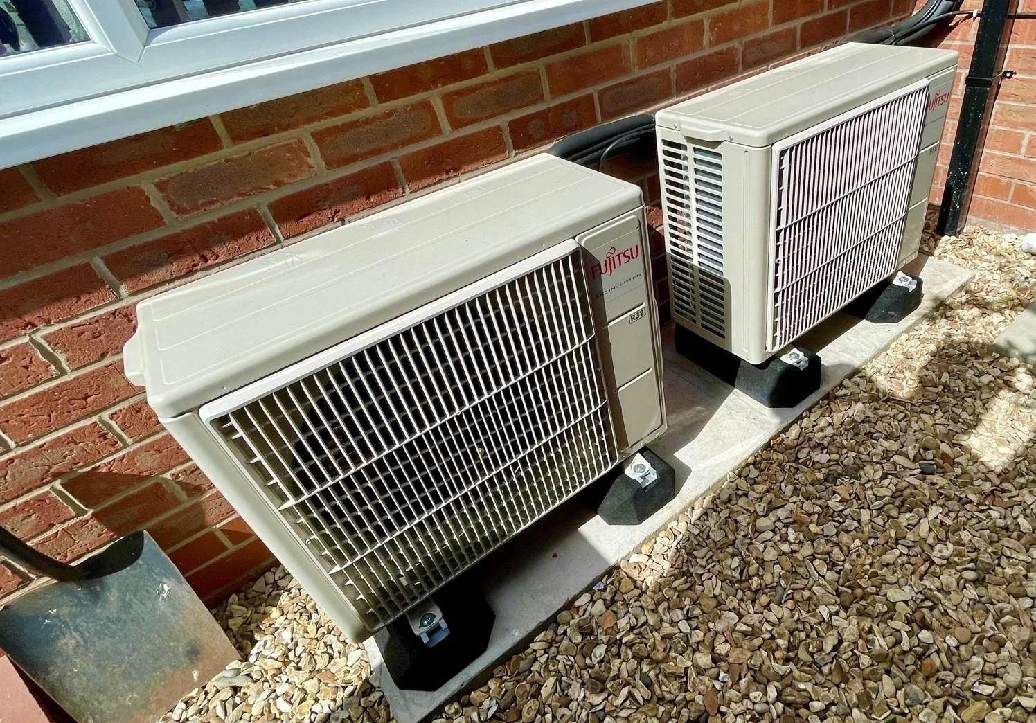 Two Fujitsu window air conditioning units installed outside a building on a concrete slab, with brick wall and gravel ground nearby.
