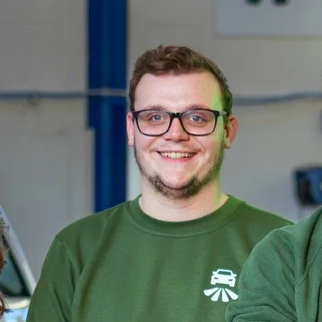 A man with curly dark hair, glasses, and a beard, wearing a dark green sweater, smiling softly in a room with a light-colored wall and a green plant in the background.