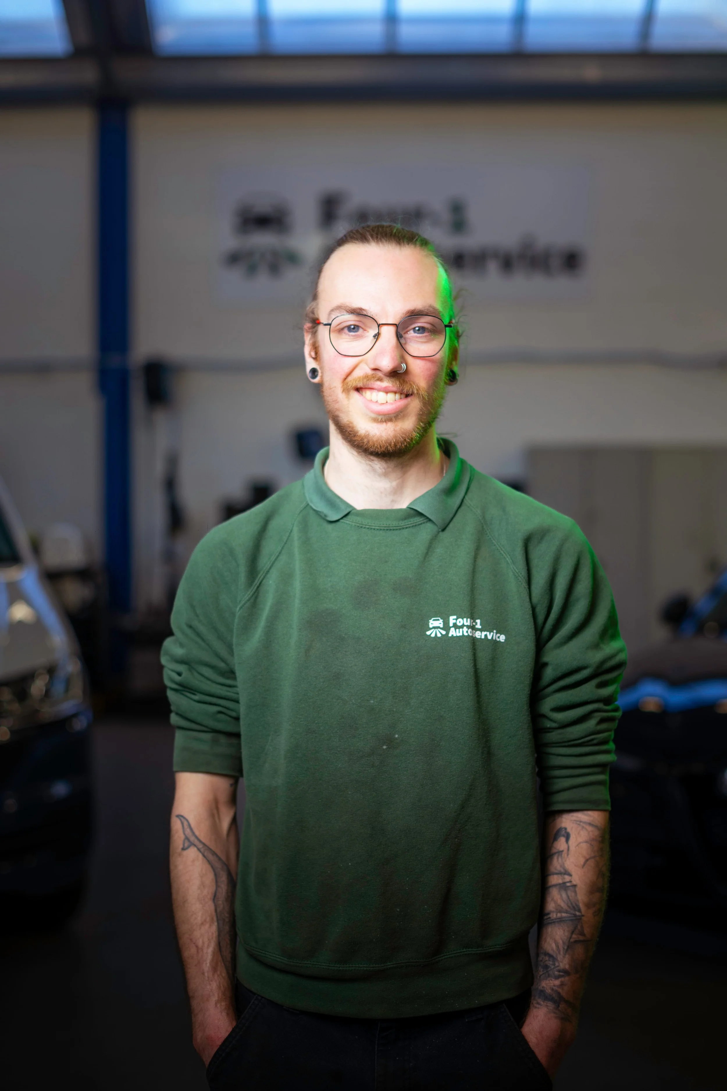 A smiling woman with curly hair wearing glasses, hoop earrings, and layered necklaces in an indoor setting.