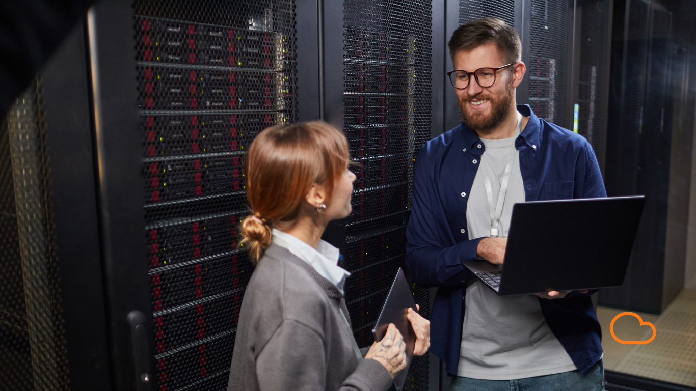 Two IT professionals, a woman with red hair and a man with glasses and a beard, standing in a server room and smiling while having a conversation.