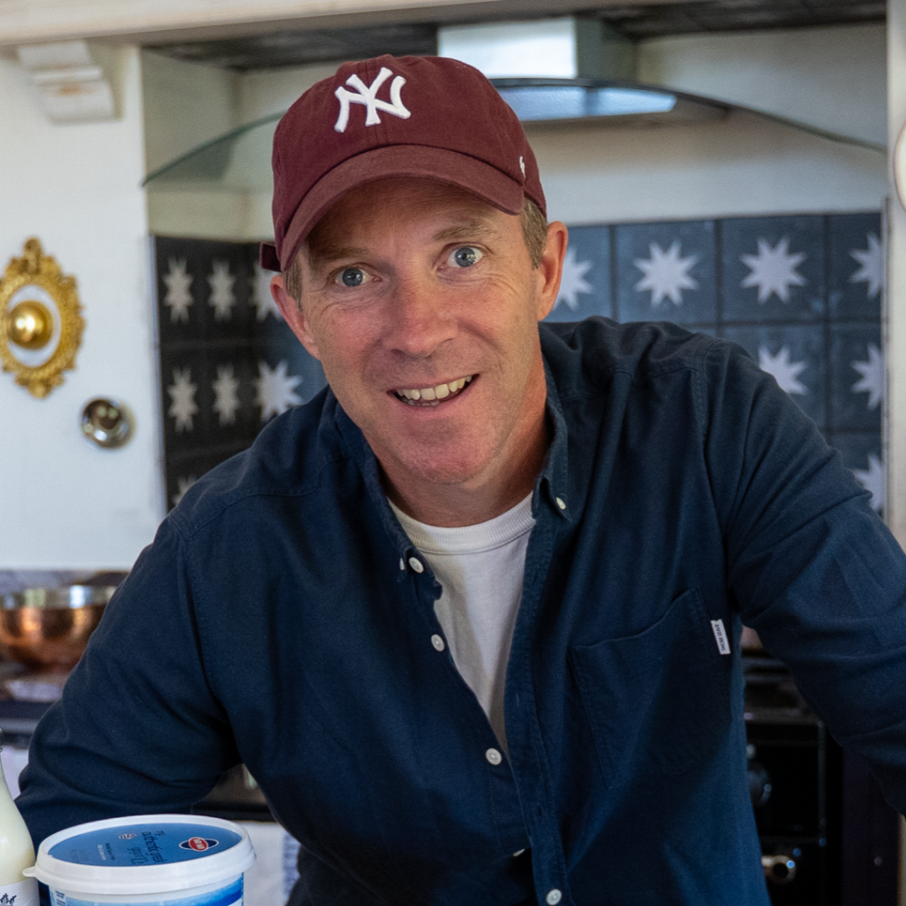A smiling man wearing a maroon New York Yankees baseball cap and a dark blue button-up shirt in a kitchen.