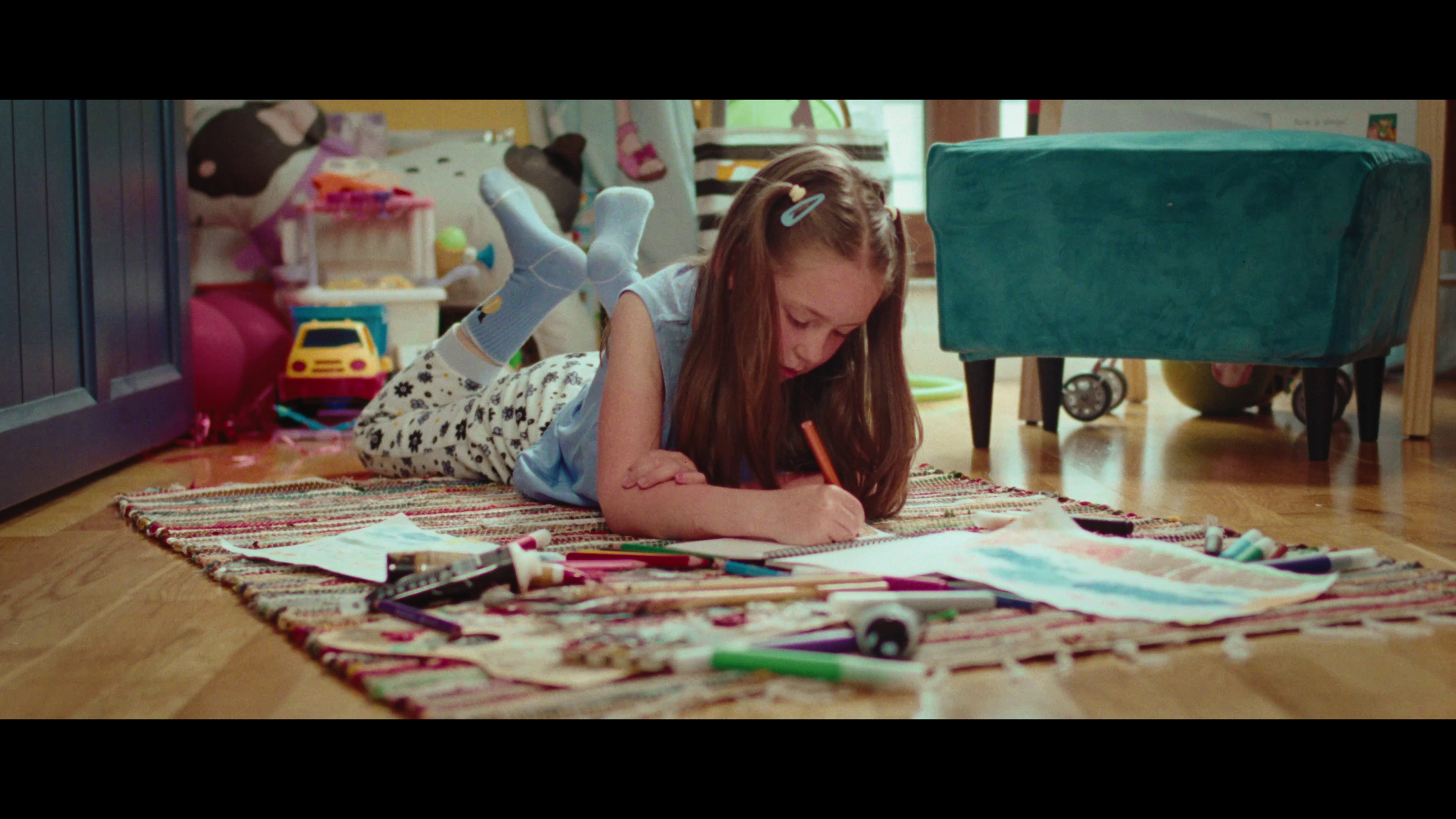A young girl with long hair lying on her stomach on a colorful rug, drawing with markers and pens, surrounded by art supplies on the floor, in a cluttered room with toys and furniture.