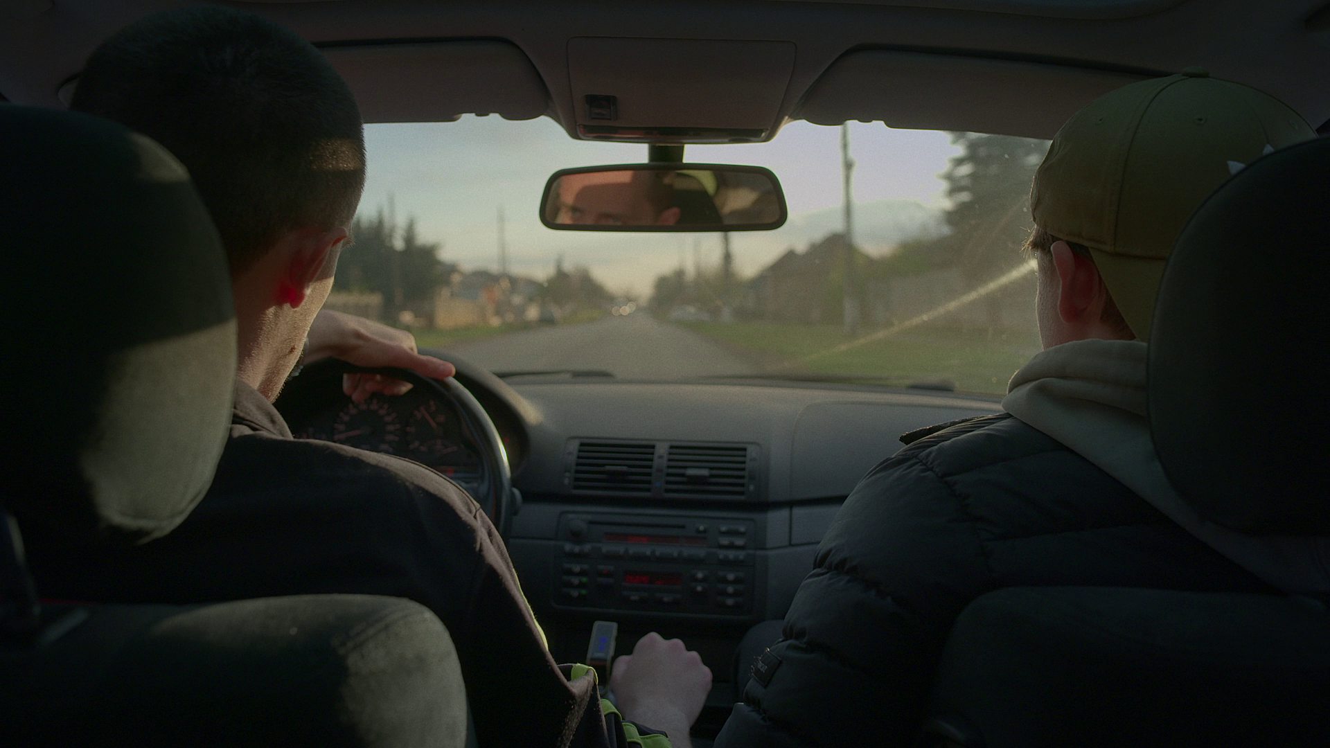 Two young men in a car, one driving and the other sitting in the passenger seat, during daytime with a view of the road ahead.