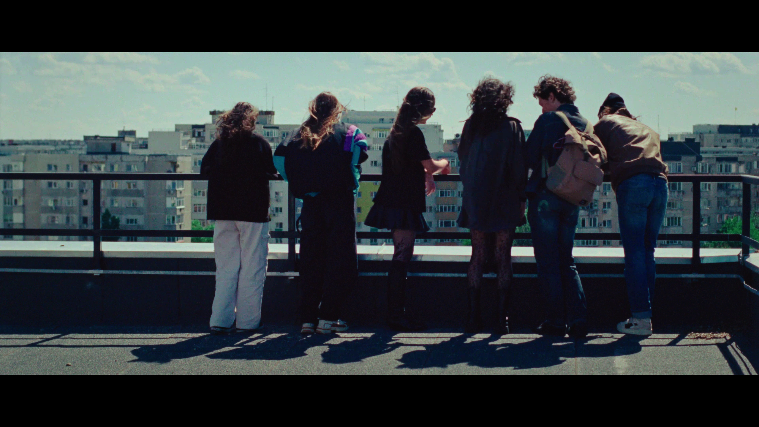 Six young people on a rooftop overlooking city buildings, standing in a row, facing away.
