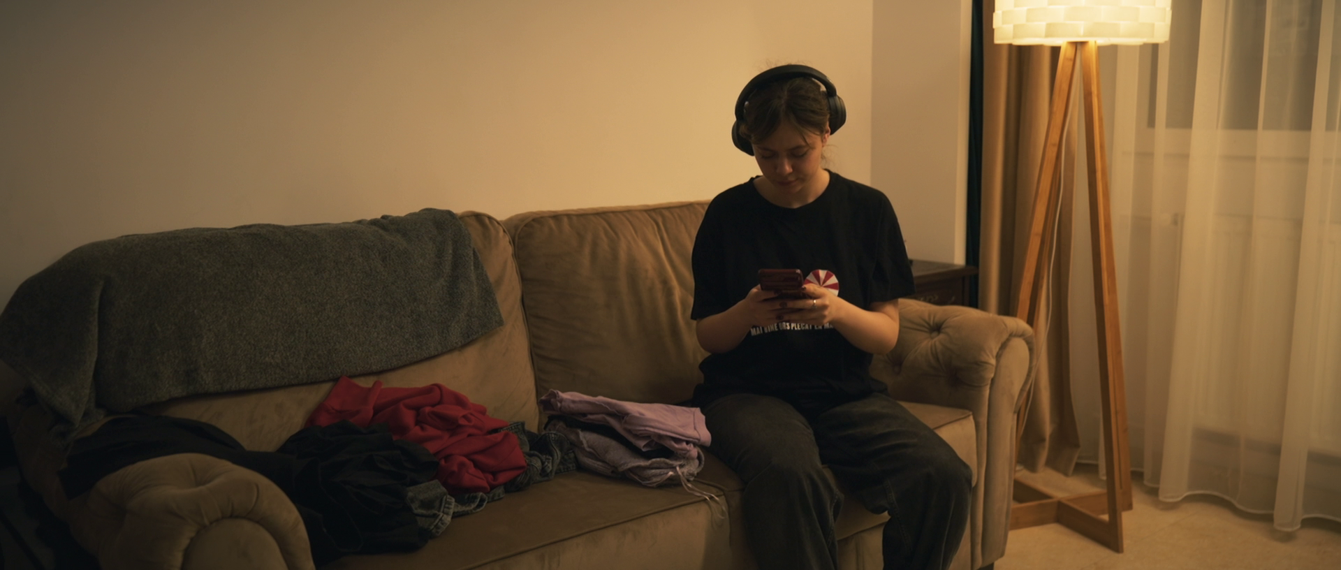 A young person sitting on a beige sofa wearing headphones and looking at a smartphone, with clothes and a blanket on the sofa, in a dimly lit room with a tall floor lamp and a window with sheer curtains.