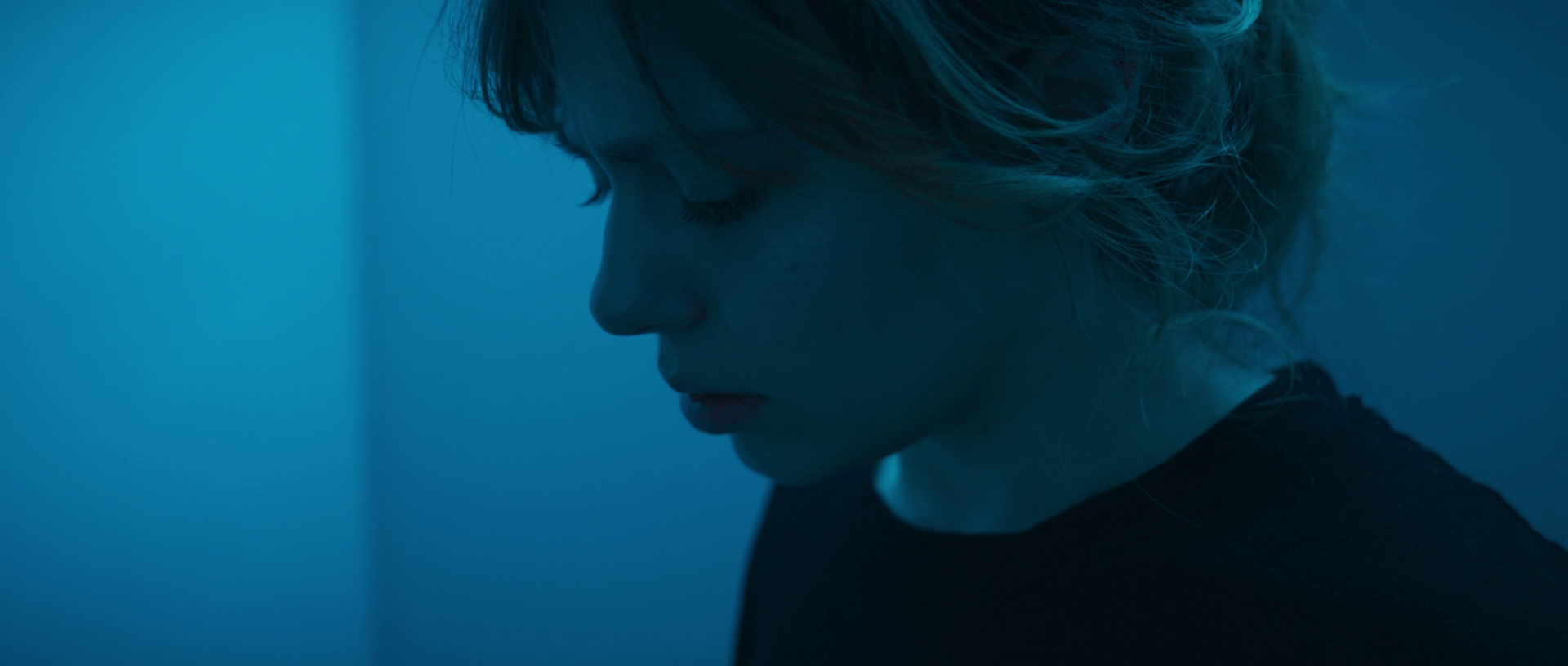 Close-up of a woman with curly hair, looking down, with a somber or reflective expression, in a dimly lit setting with blue tones.