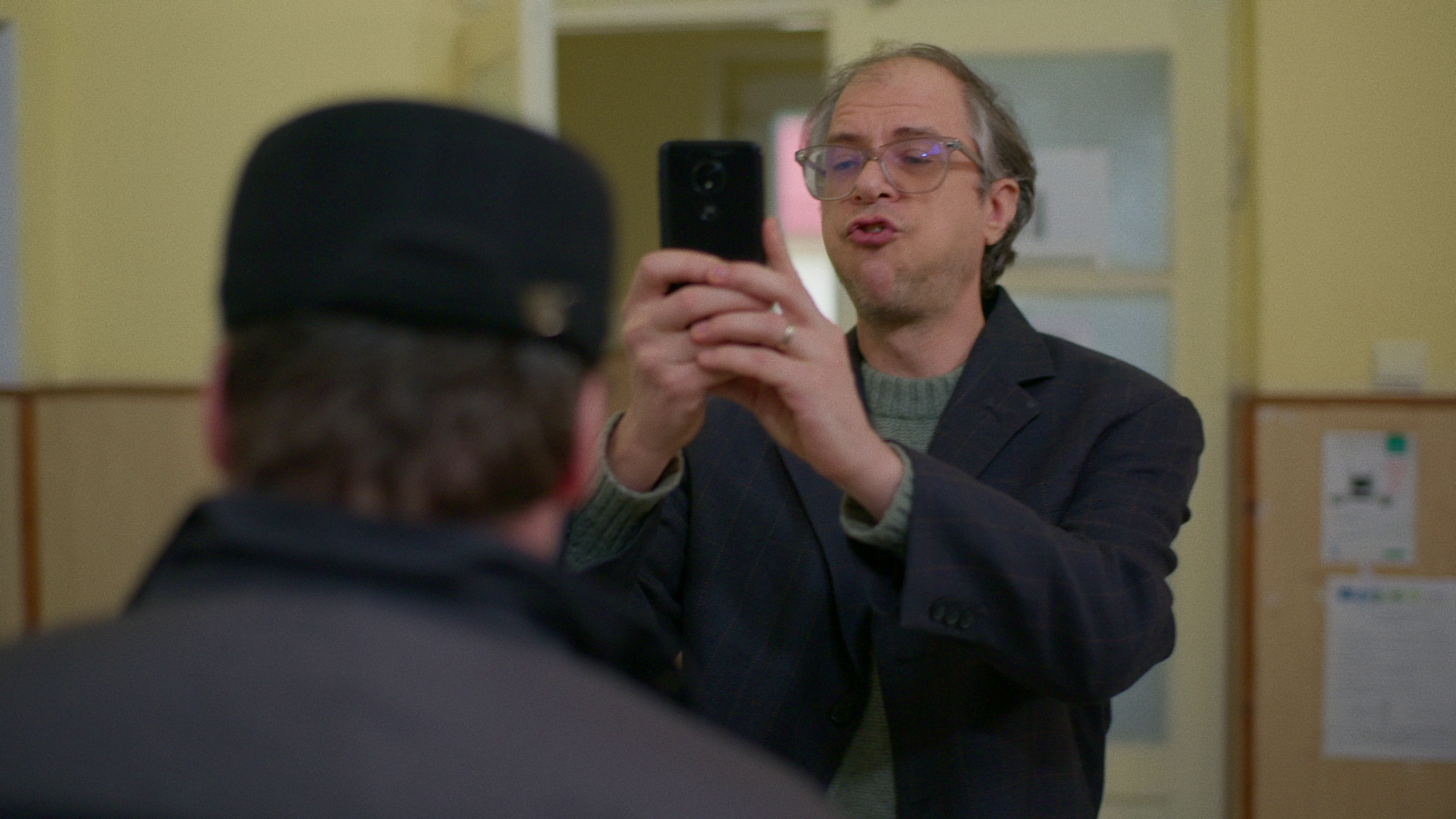 A man with glasses and gray hair is taking a photo with his phone of another person wearing a black hat and dark clothing in an indoor setting with yellow walls and bulletin boards.