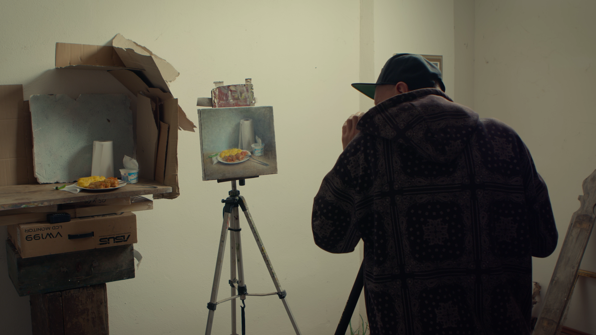 A person wearing a black hoodie and cap is taking a photo of a still life painting on an easel in a room with plain white walls. The artwork depicts a plate of food, a cup, a fork, and a yogurt container.