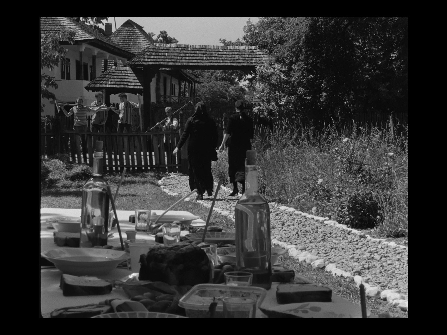 Black and white photo of people walking on a garden path near a table with drinks and food, with a playground and houses in the background.