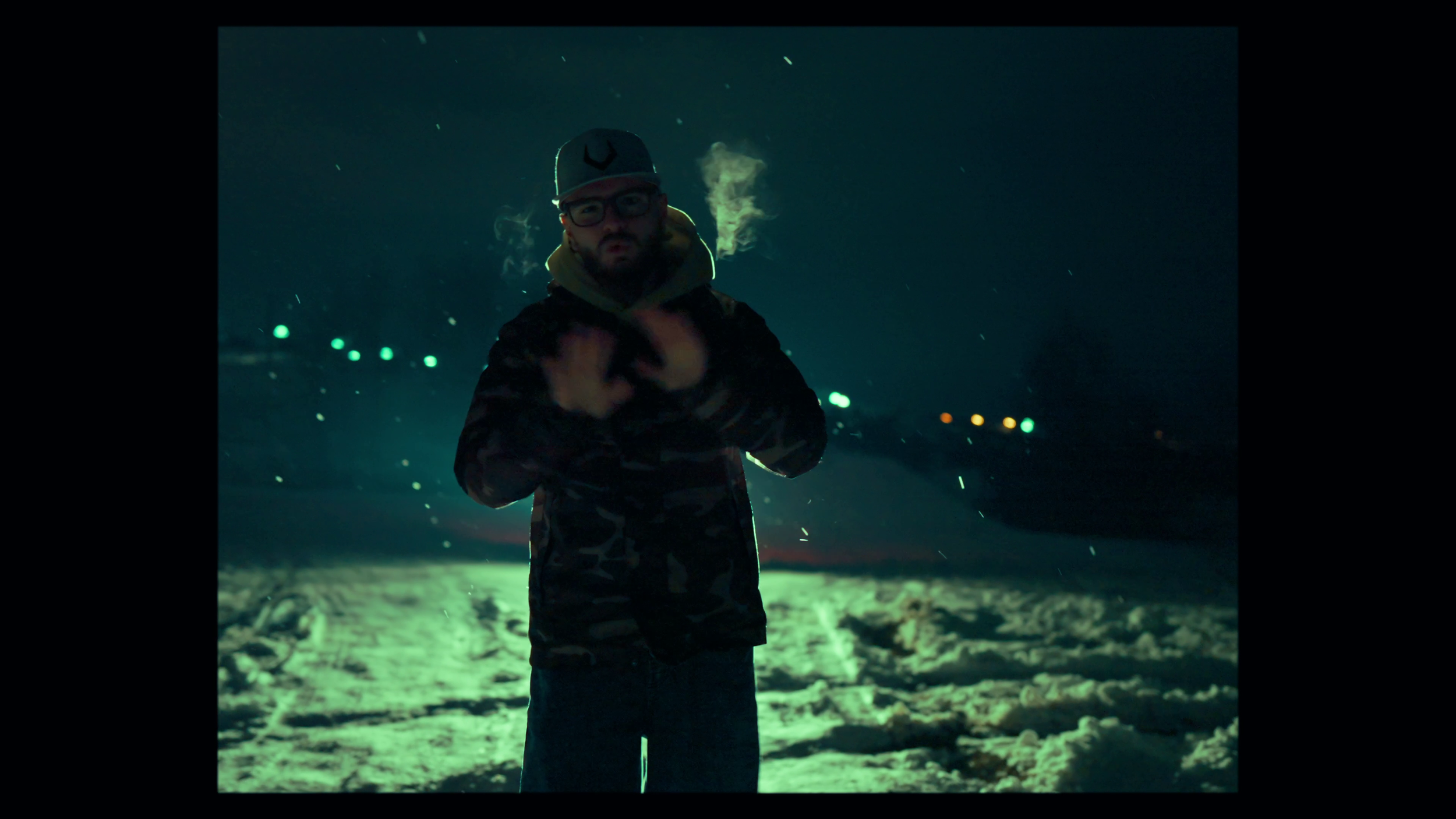 Man standing outdoors at night in snow, wearing a cap and camo jacket, with steam near his face and colorful lights in the background.