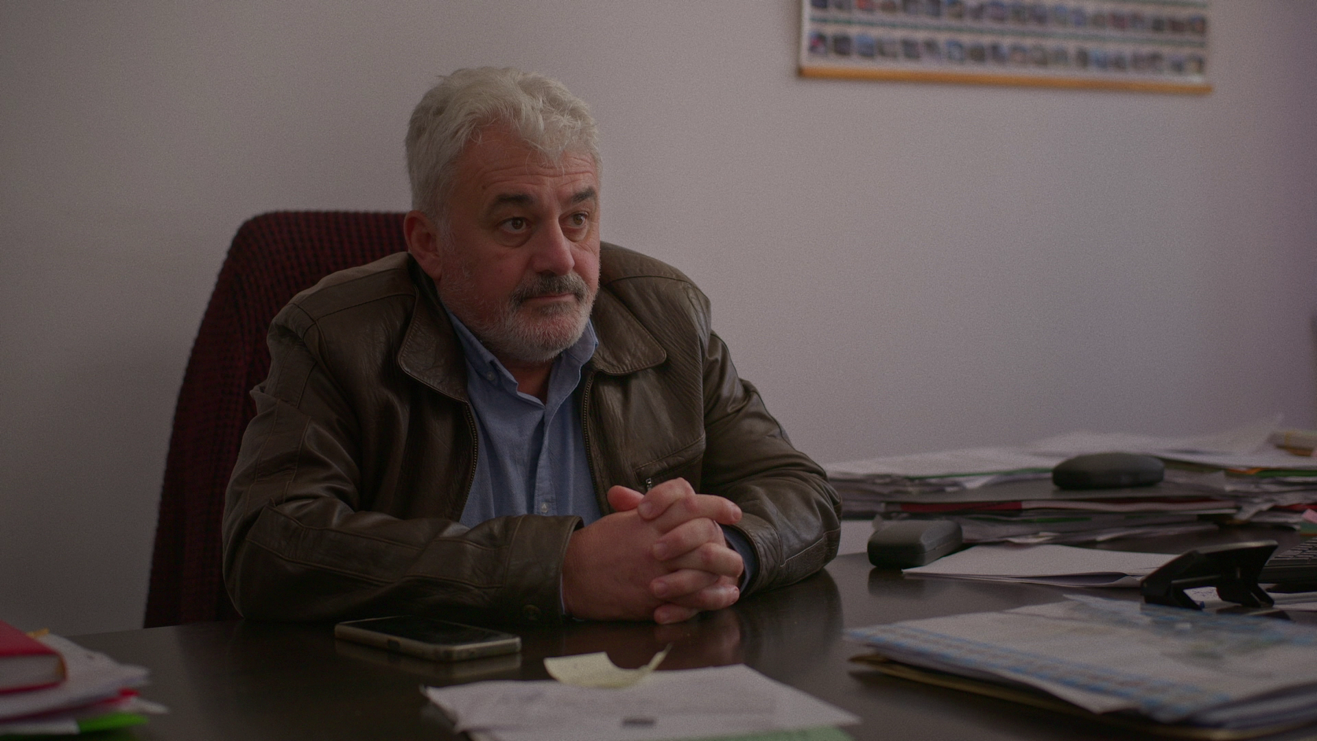 An older man with gray hair and a beard sitting at a cluttered desk, wearing a brown leather jacket and a light blue shirt, with his hands clasped in front of him, looking serious or contemplative.