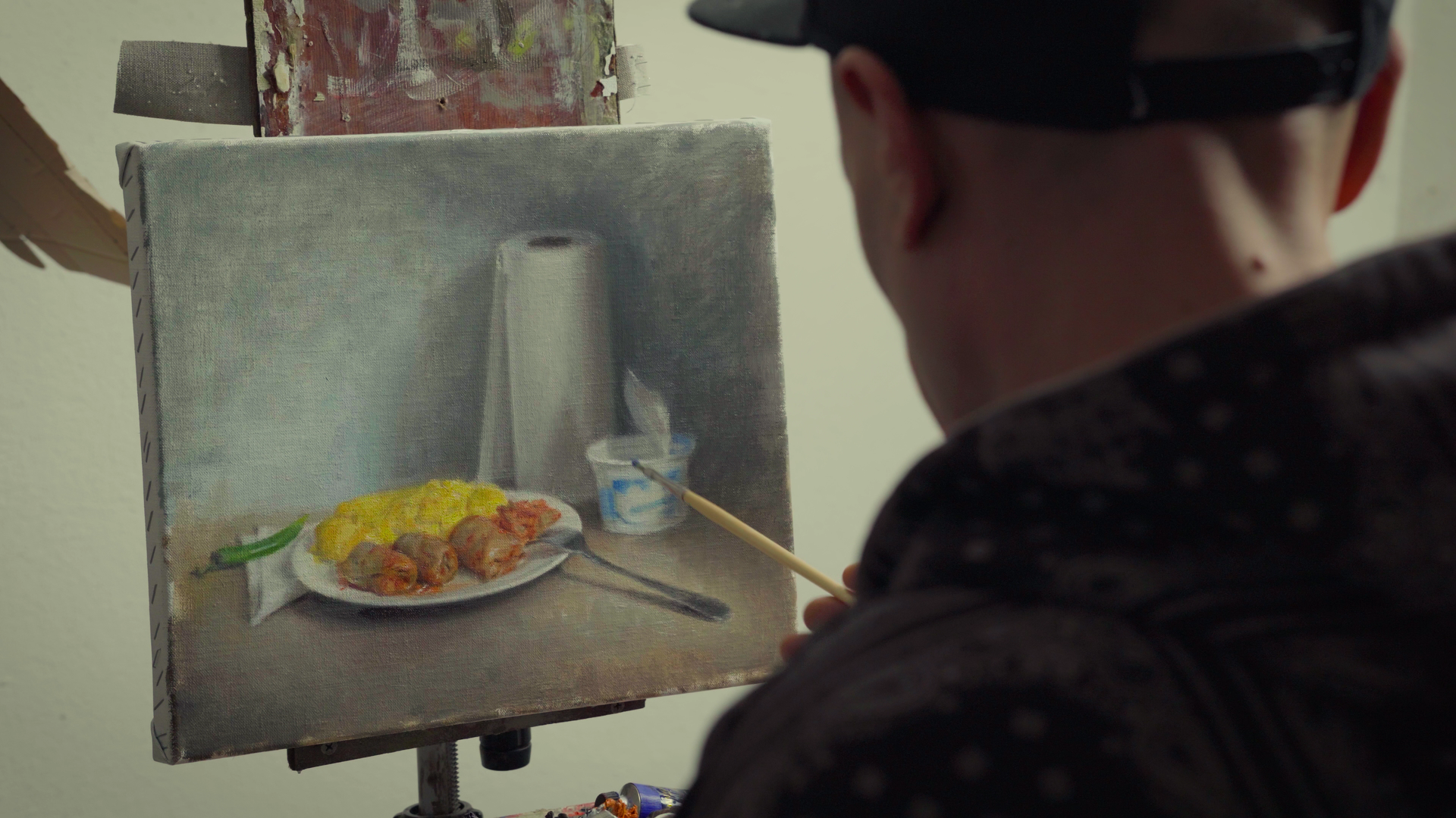 An artist painting a still life scene of a plate of food, a salt shaker, and a small container on a canvas.