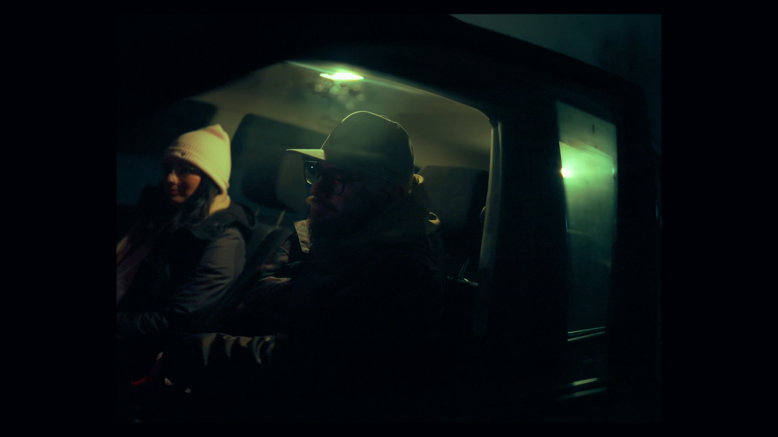 Two people sitting inside a vehicle at night, illuminated by faint greenish light, one wearing a white beanie and glasses, the other wearing a baseball cap and glasses.