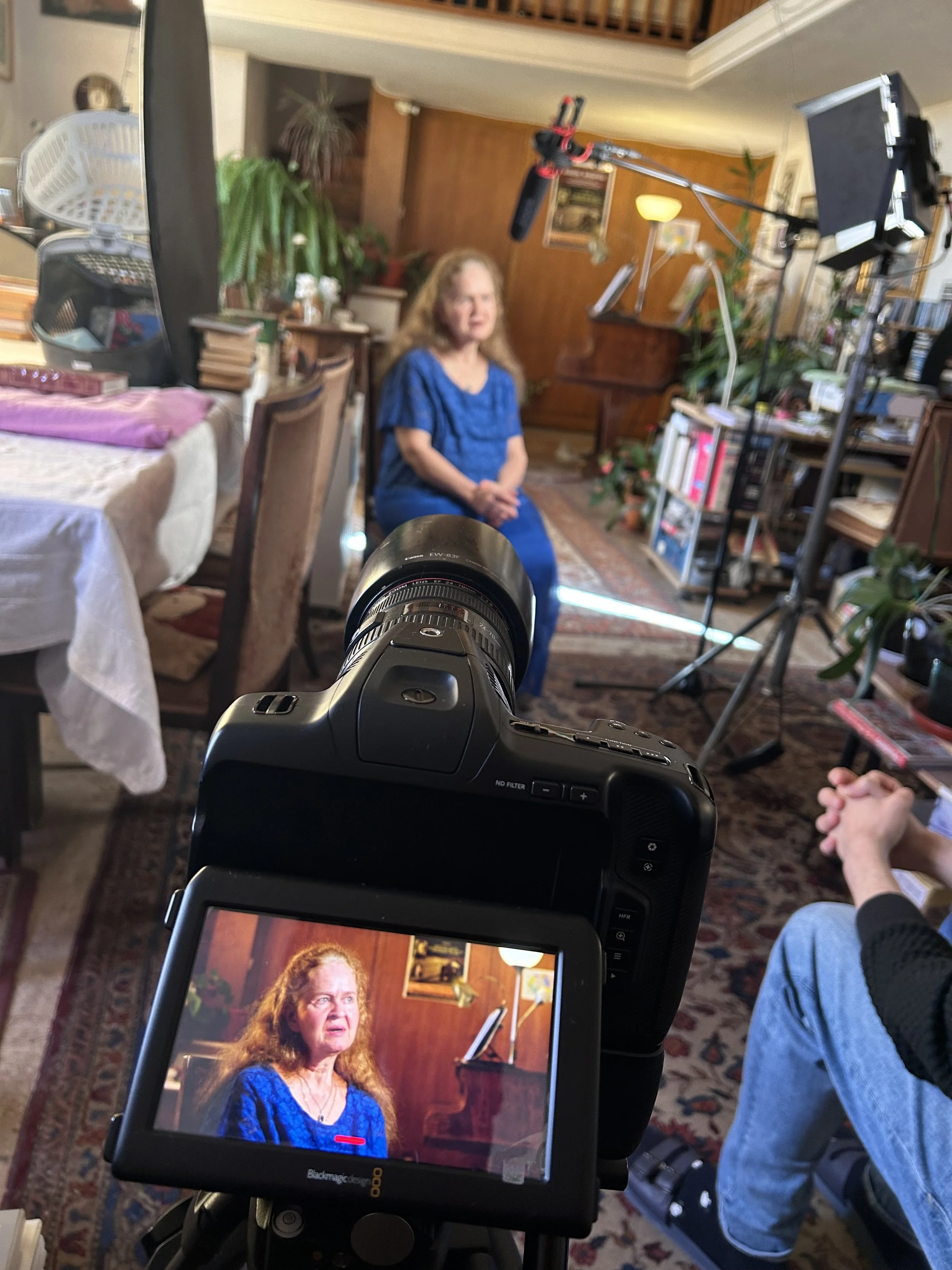 A woman with long curly hair wearing a blue shirt, sitting in a room with wooden walls, plants, and furniture, being filmed for an interview with professional lighting and camera equipment.