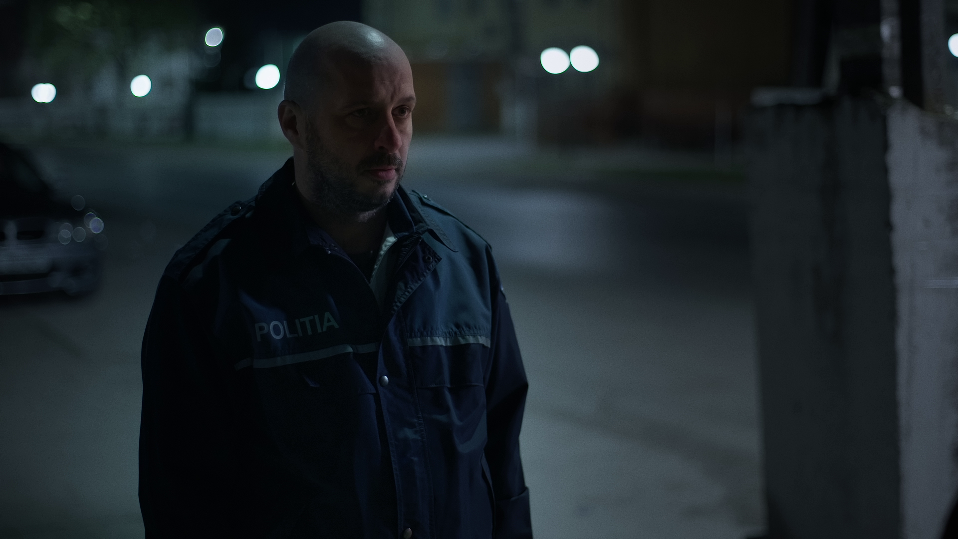 A police officer standing alone at night in a dimly lit outdoor setting, with a serious expression.
