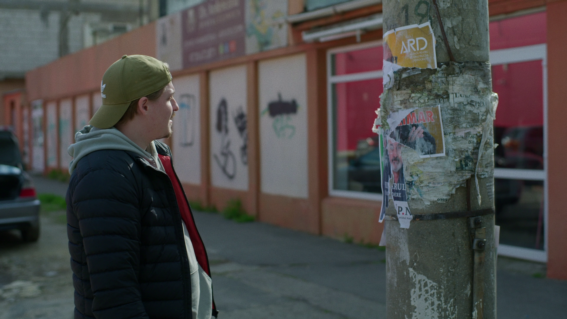 A young man wearing a black puffer jacket, gray hoodie, and khaki cap looks at a weathered utility pole with torn posters. The background features a red-orange building with large windows and parked cars.
