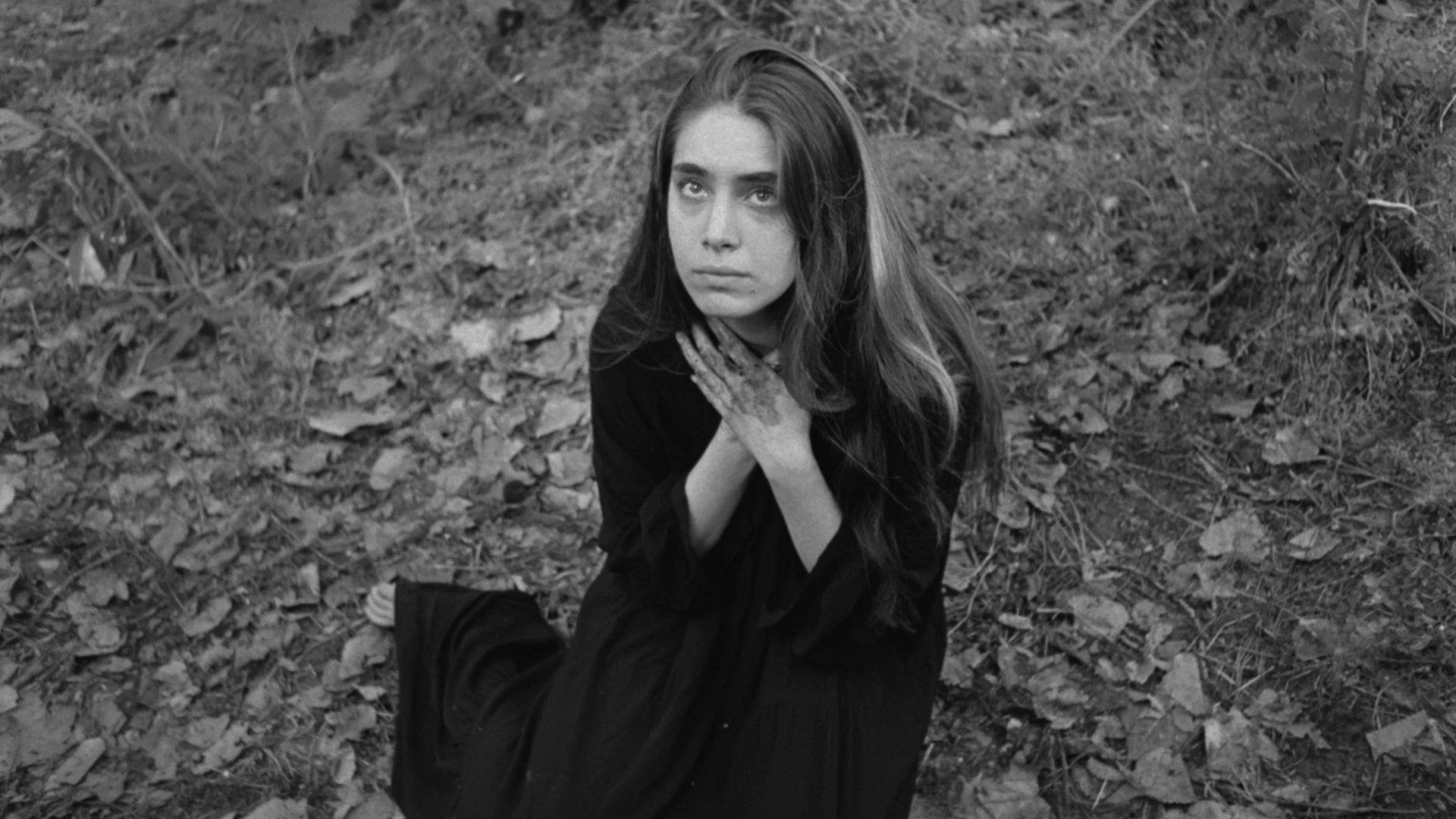 A woman with long hair, sitting on the ground in a forest, looking up, wearing a dark dress, with her hands on her chest, surrounded by leaves and natural debris.