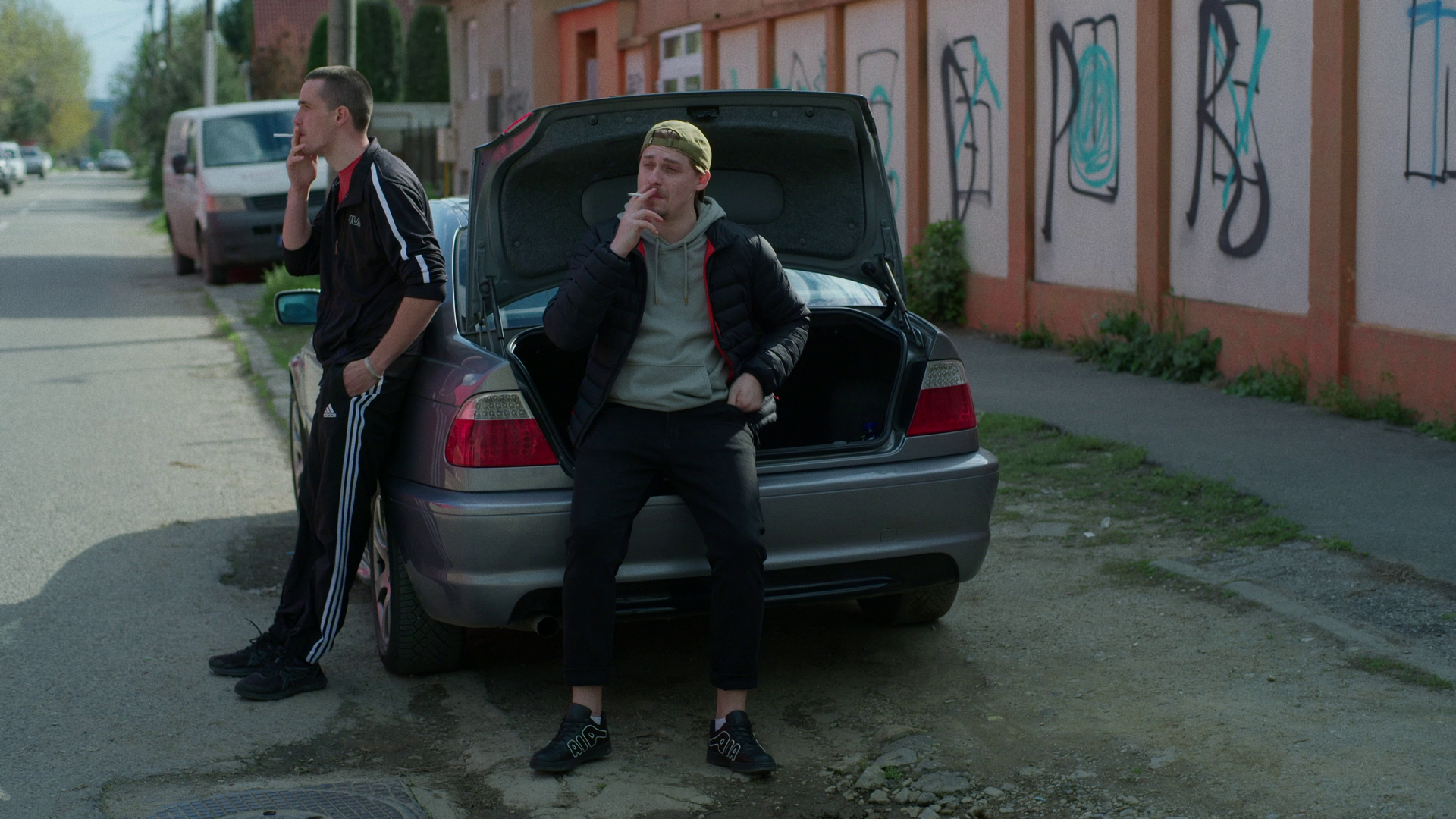 Two young men with close-cropped hair, wearing casual clothing, are standing outside near a gray car with its trunk open. One man is leaning against the car, looking away, while the other is sitting on the edge of the trunk, smoking a cigarette and looking slightly tired or contemplative. The scene is in an urban area with a graffiti-covered wall and a sidewalk.