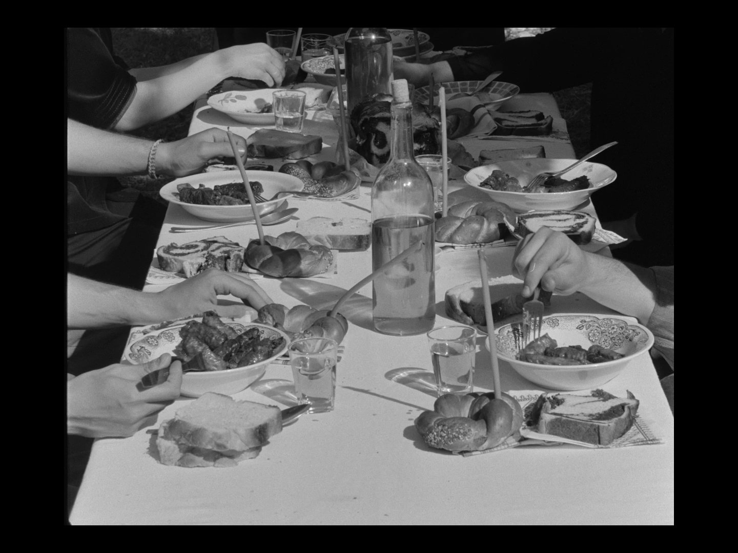 A black and white photo of a group of people having a meal outdoors at a long table filled with plates of sandwiches, bowls of food, bread, drinks, and some fruits, with their hands reaching for food.