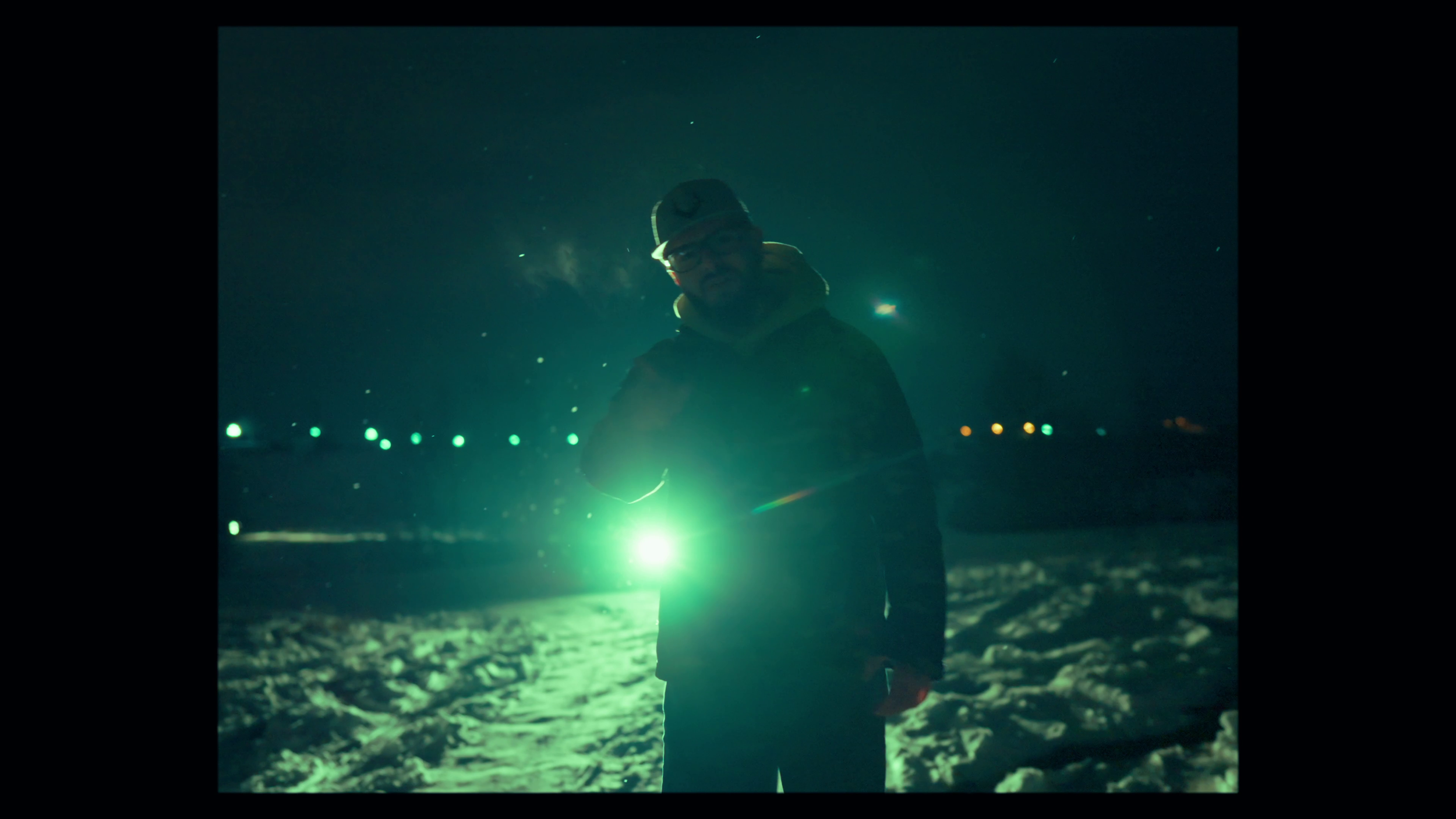 Person standing in a snowy landscape at night, holding a flashlight that illuminates their face and the snow around them.