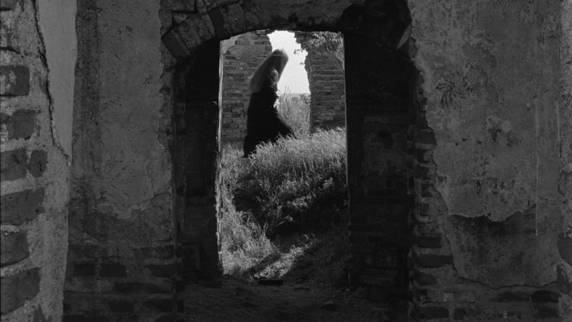 A woman in a black dress walking through an arched doorway of an old, weathered stone or brick building, with grass and trees visible outside.