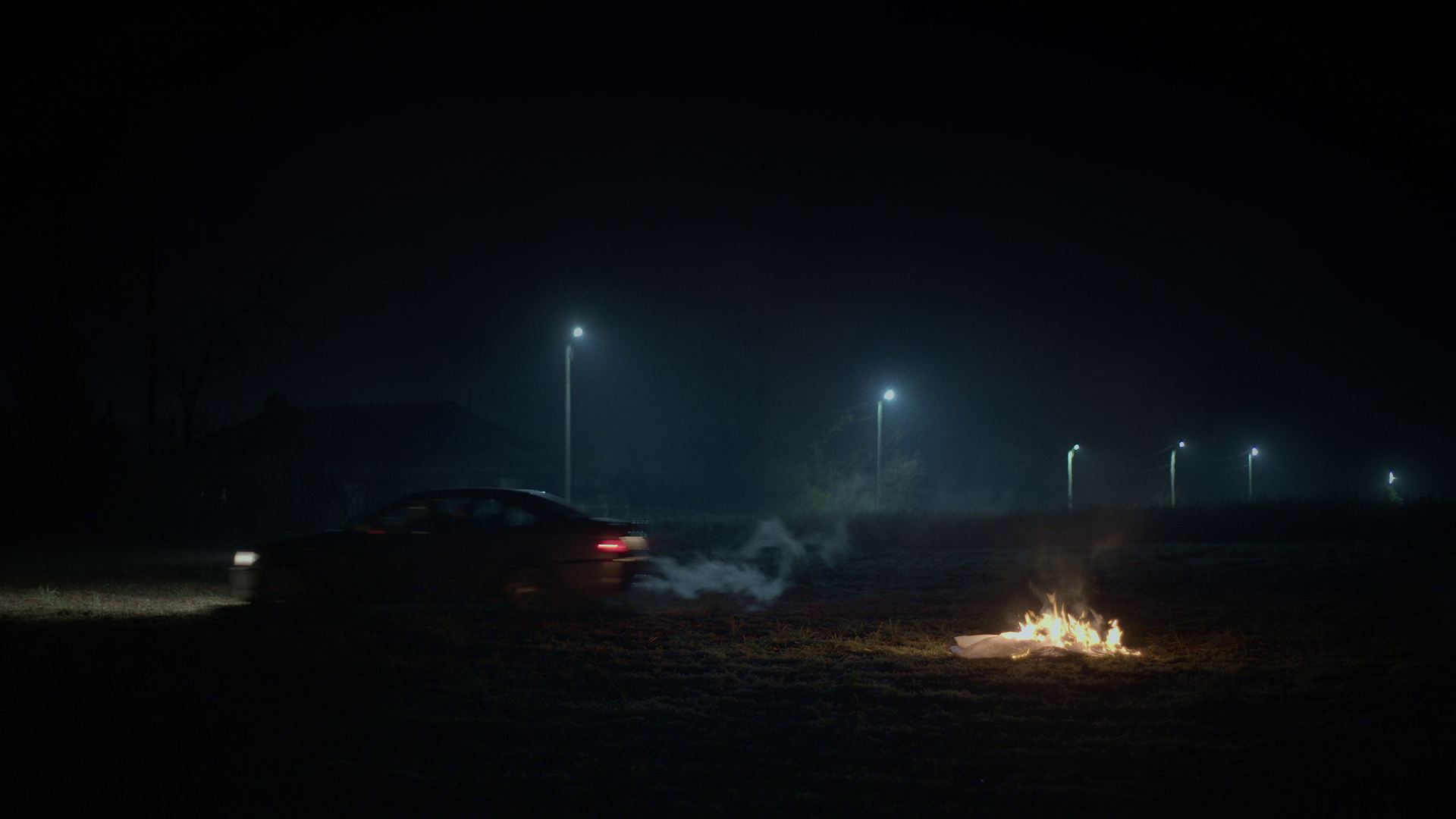 A dark outdoor scene at night with a burning object on the ground in front, a car driving by creating smoke, and streetlights in the background.