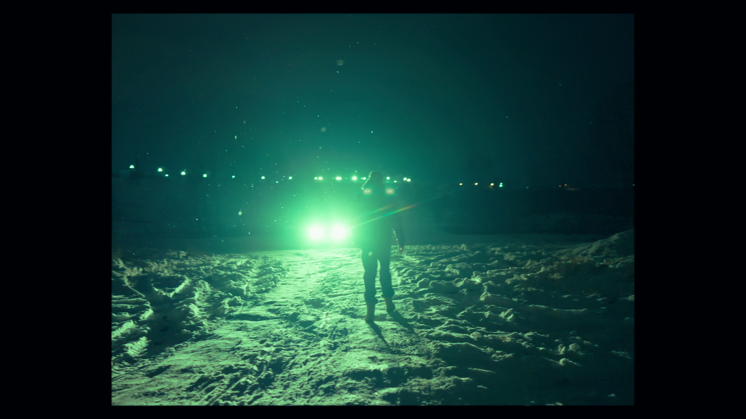 A person standing on snow-covered ground at night, illuminated by bright green lights from a vehicle or equipment, with a dark sky and distant lights in the background.