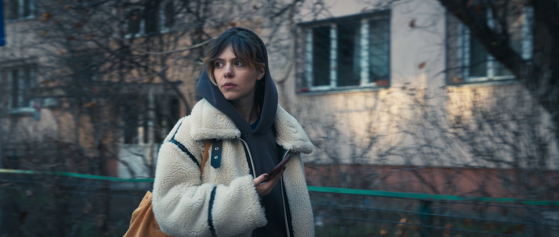 Young woman with short brown hair and light skin wearing a black hoodie and tan shearling jacket walking outdoors while looking to her left and holding a smartphone.