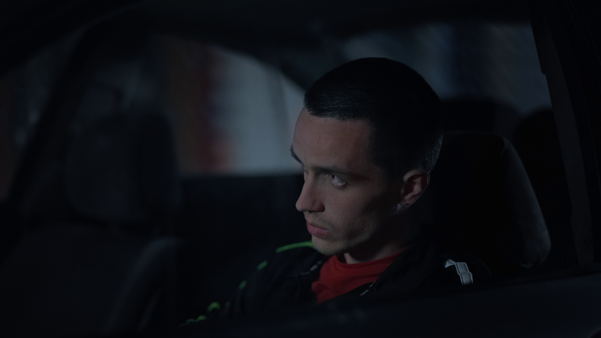 Young man with short dark hair sitting in a dark car, looking out the window with a serious expression.