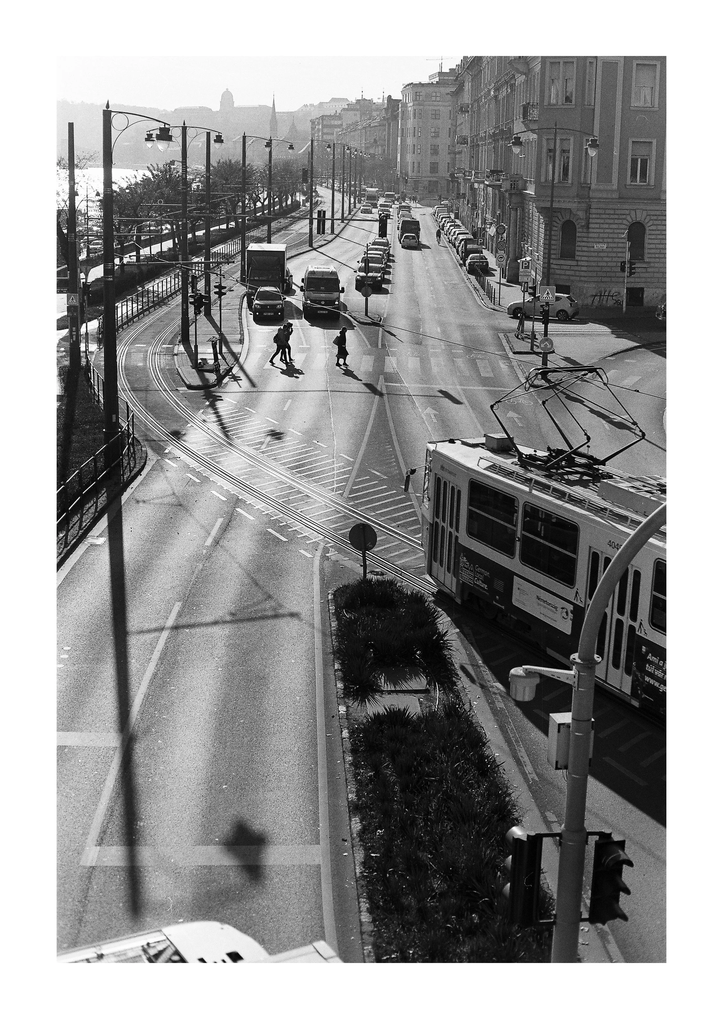 Black and white photo of city street with tram, cars, pedestrians crossing, and buildings in background.