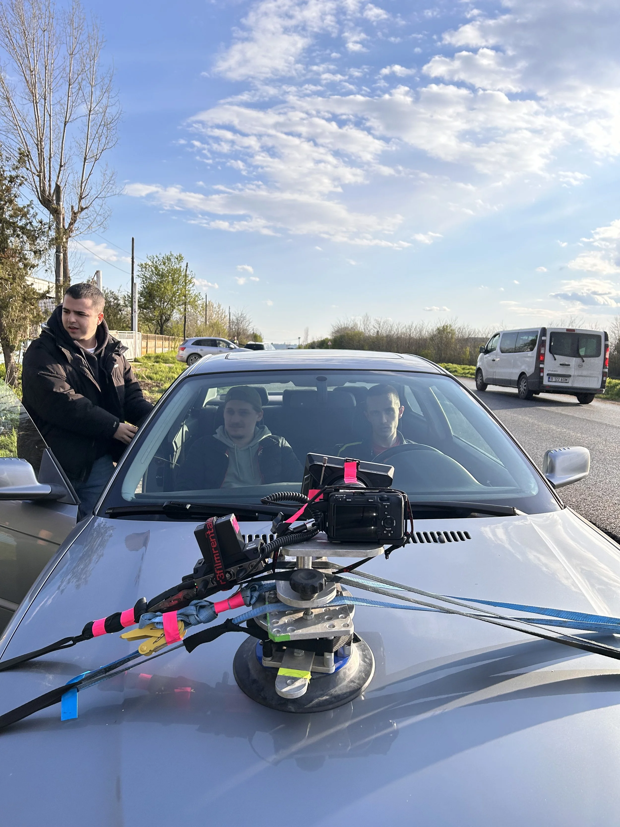 Three men on a road with a camera mounted on the hood of a silver car, used for filming or surveying, with other vehicles and a rural landscape in the background.