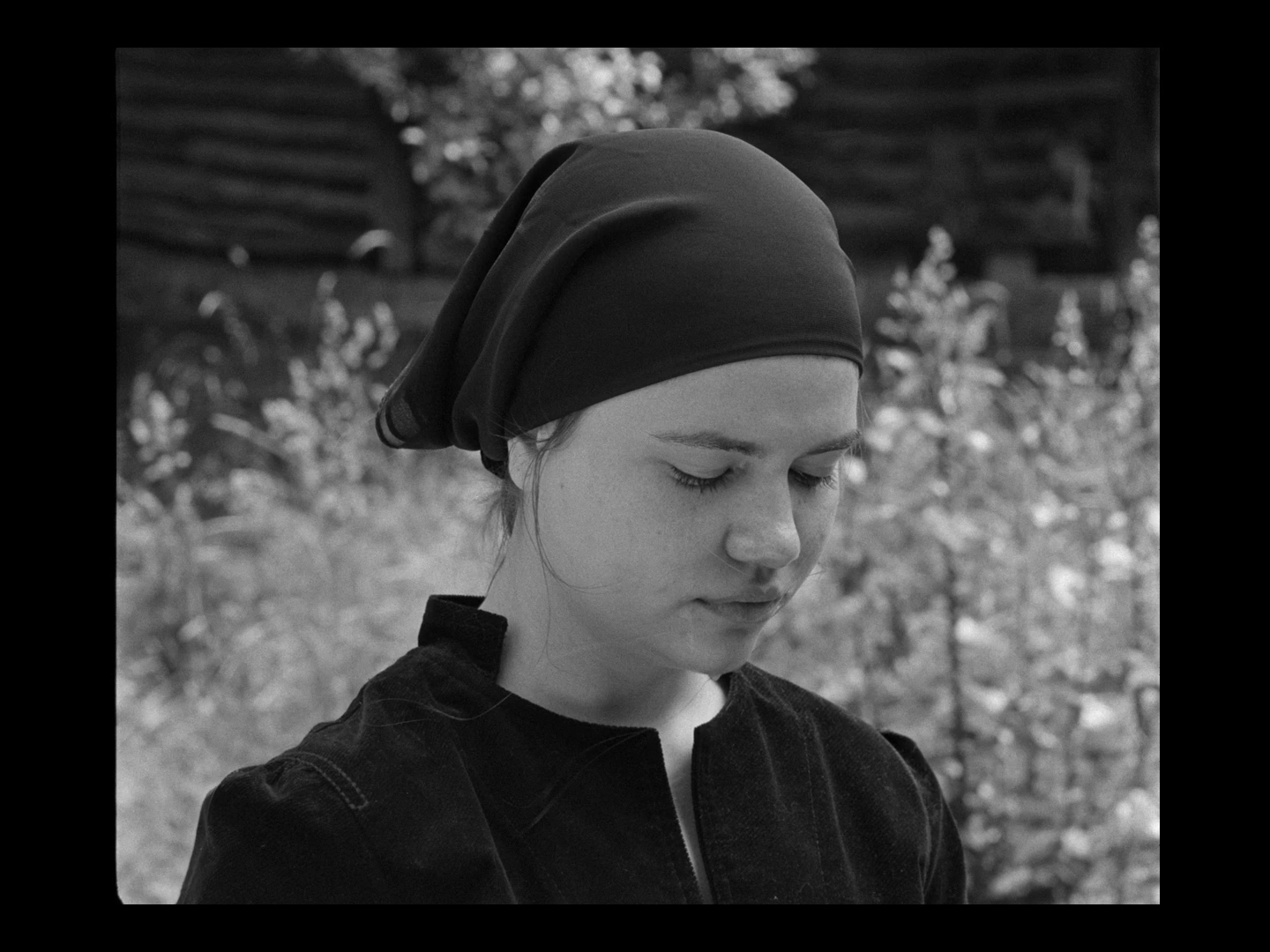 Black and white photo of a young woman with her eyes closed, wearing a headscarf and a jacket, outdoors with foliage and a wooden fence in the background.