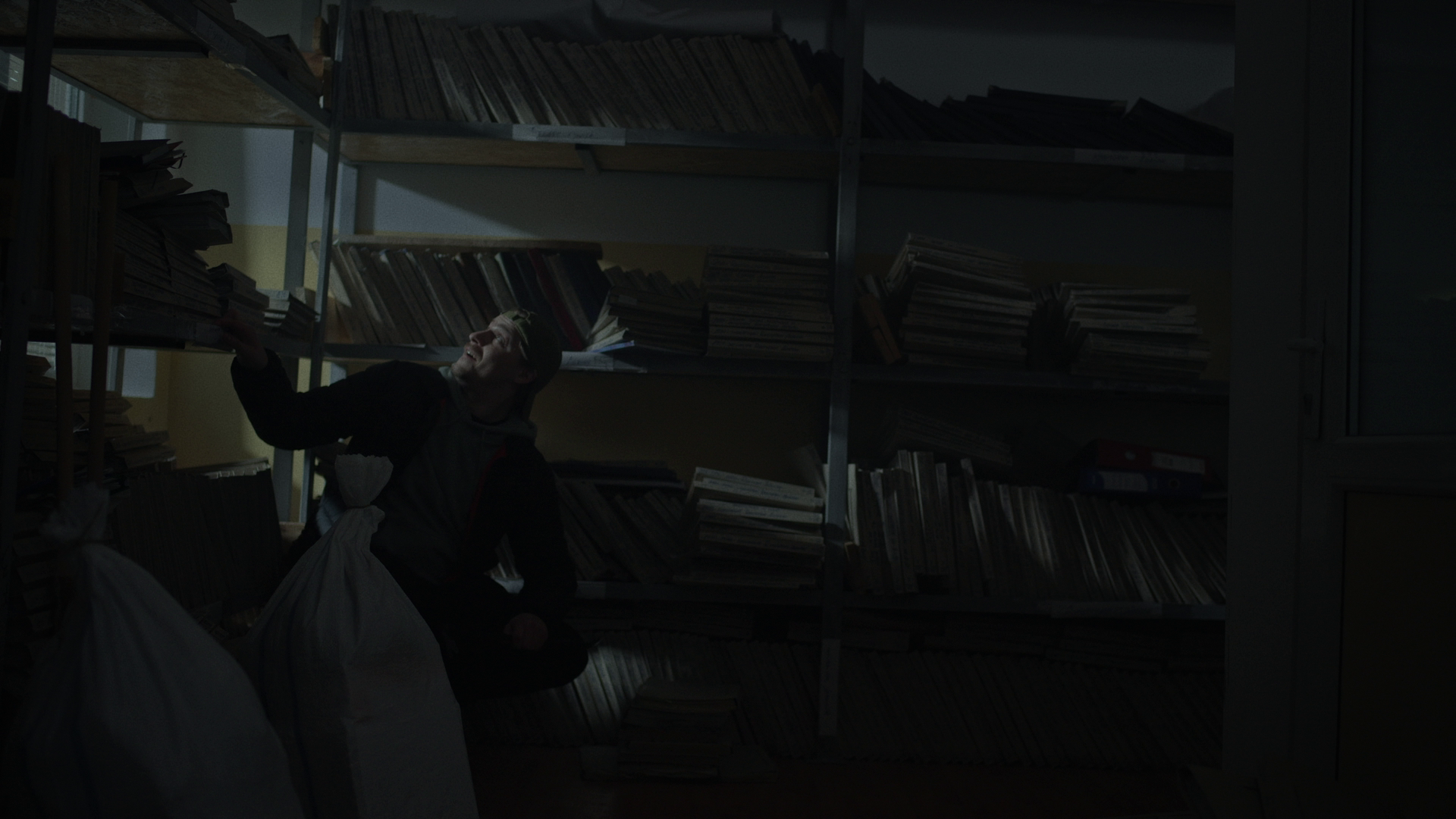 A person inside a dimly lit room with shelves filled with papers and folders, reaching for a folder on a high shelf.