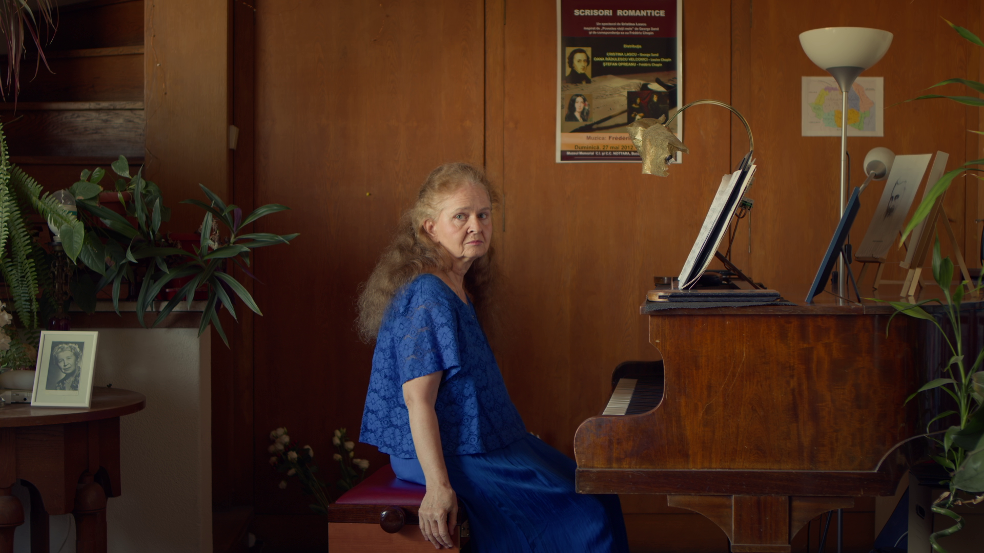 An elderly woman with long, curly, gray hair sitting at a wooden piano in a room with wood-paneled walls and various plants. She is wearing a blue dress and looking towards the camera with a serious expression.