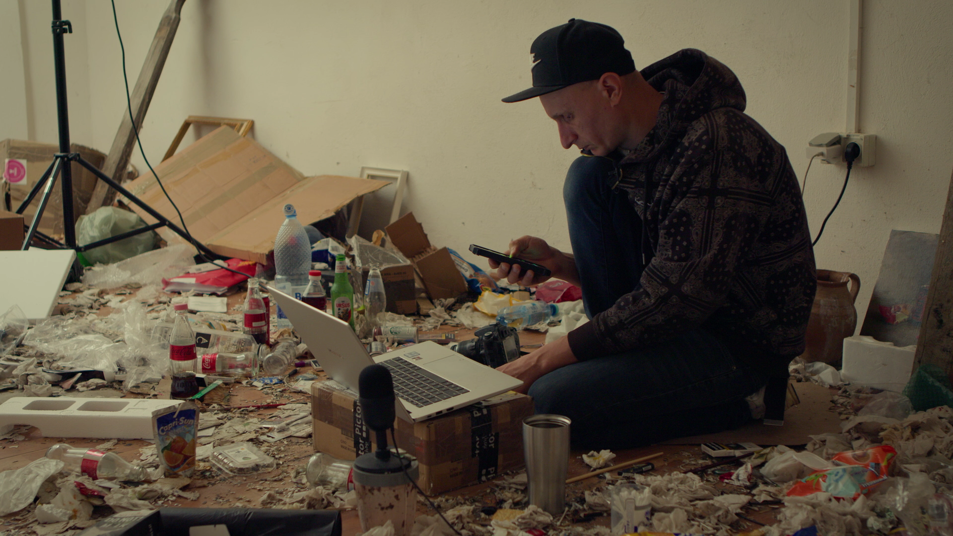 Young man sitting on the floor in a messy room surrounded by trash, bottles, and electronics, using his phone and with a MacBook laptop and a camera nearby.